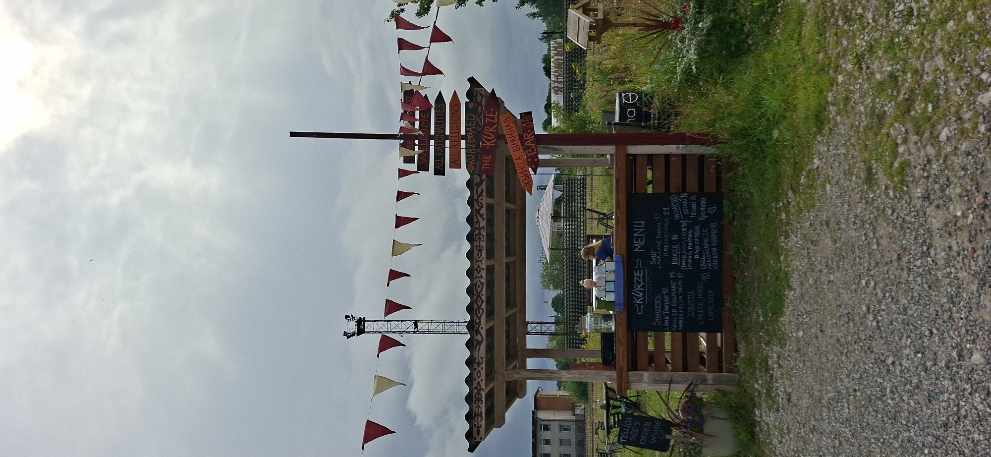 The image depicts an outdoor food stand in Tallinn, Estonia. The stand, constructed from wood, features a covered roof adorned with decorative patterns. A menu board is prominently displayed, listing various food items and their prices. Among the offerings are "Starters" like Lamb Tartar and Grilled Eggplant, as well as "Chutty" options such as Cheese, Herbs, Lamb, and Beef. The menu also includes "Kurze" items and "Chiken Kebab".
Several directional signs are mounted on a pole near the stand, with text that appears to include "ARE WELCOME," "ALL NATIONALITIES," "ALL RELIGIONS," and possibly place names or other information in a language other than English. One sign explicitly reads "THE KURZE".
Two individuals are visible behind the counter of the food stand. One person, with blonde hair pulled back, appears to be serving or preparing food, while another individual, wearing glasses and a blue shirt, is also present. Several mugs are stacked on the counter.
In the background, a tall transmission tower with lights is visible, along with a series of red and yellow triangular flags strung across the scene. The environment is grassy with some vegetation, and a gravel path leads up to the food stand. A birdhouse is mounted on a post to the right of the stand. The sky is overcast, suggesting a cloudy day.