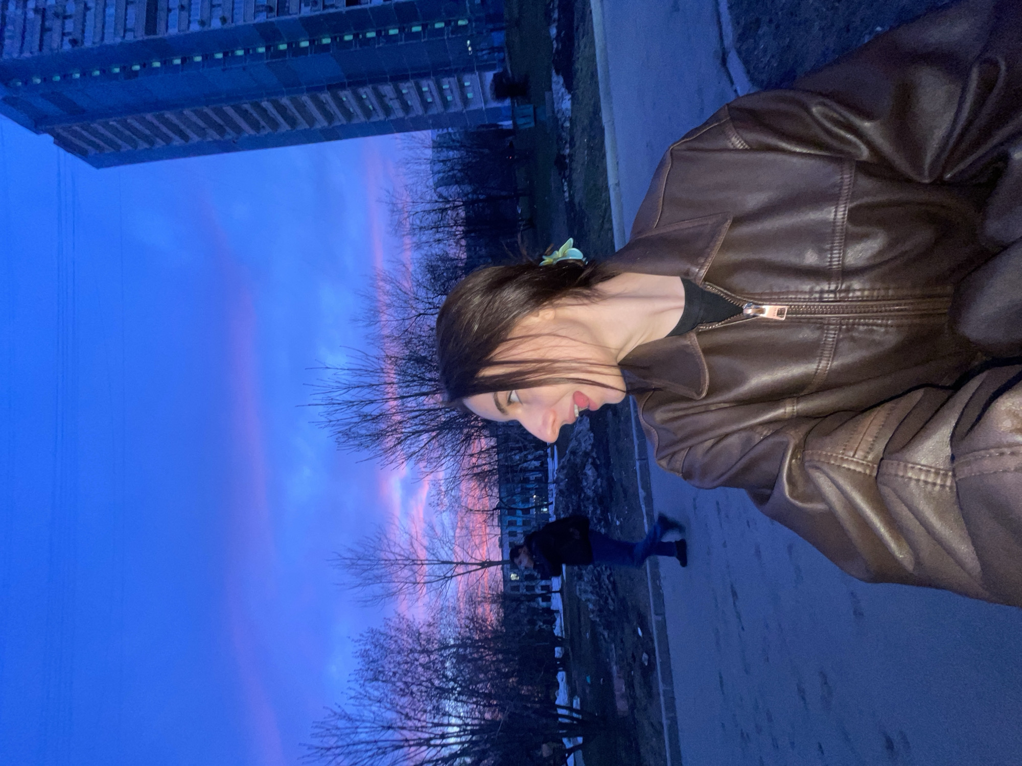 The image captures an urban outdoor scene at twilight. In the foreground, positioned to the right, a young female individual is partially visible from the chest up, her head turned sharply to her right (viewer's left). She has dark hair, styled with a light-colored, possibly green or blue, hair accessory. She wears a brown, zippered leather-look jacket over a black garment. Her mouth is open. In the mid-ground, a male individual walks from left to right on a paved path, wearing a dark jacket and dark trousers. He holds a dark object to his right ear, consistent with a mobile phone.
The background features numerous bare deciduous trees, indicating a non-summer season. A tall, multi-story residential building, characteristic of a *panelka*-style apartment block, stands prominently on the far right, displaying several illuminated windows with a greenish hue. Another, darker building is partially obscured by trees on the left. The sky exhibits a deep blue overhead, transitioning to distinct pink and purple hues along the horizon. The ground consists of paved areas and unkempt grassy or dirt patches.
The architectural style, specifically the *panelka* apartment building, and the general urban environment, are consistent with residential areas found in Moscow, Russia.