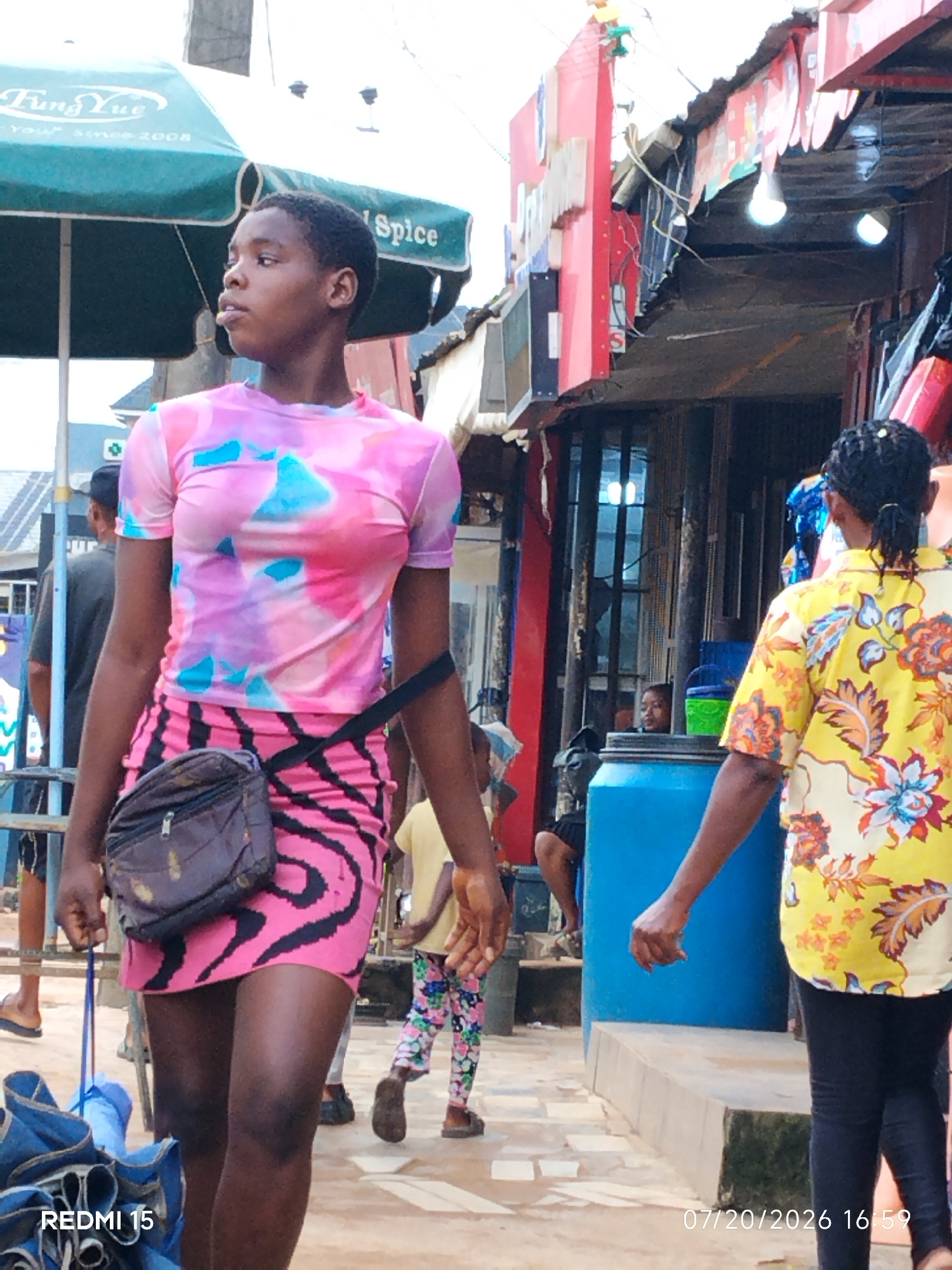 The image depicts a street scene in Umuode, Nigeria, with a focus on several individuals in a market-like environment. In the foreground, a young person wearing a pink and blue tie-dye t-shirt and a pink and black tiger-striped skirt is prominently featured, looking to the left. A dark brown crossbody bag is slung across their body. To the right, another individual is visible from the back, wearing a yellow shirt with a floral print and black leggings. Behind them, a large blue plastic container is situated on a raised concrete structure. Further in the background, other people and market stalls are visible, including a green umbrella with the branding "Fung Yue" and "Spice". The ground appears to be a mixture of paving stones and dirt. The lighting suggests it is daytime, with some areas in shadow and others illuminated. The overall scene conveys a bustling, everyday activity in a Nigerian urban setting.