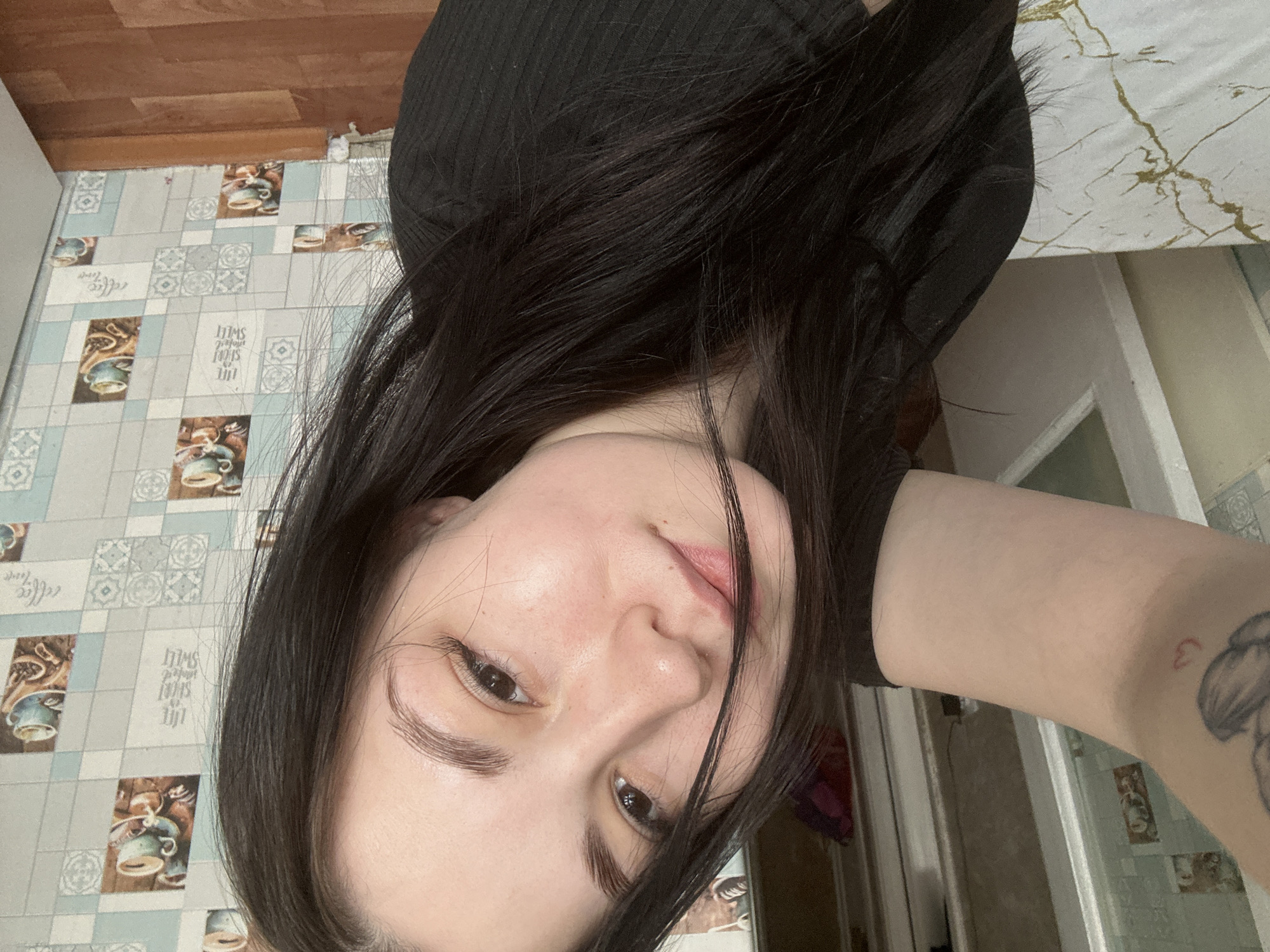 A person with dark hair is lying down, facing the camera. The person has fair skin and dark eyes. They are wearing a dark-colored garment. Part of a tattooed arm is visible on the right. The background includes a tiled floor with coffee-themed patterns, a wooden floor, a white wall, and a door. Based on the tile pattern and other clues, the location is likely in Chaikovsky, Russia.