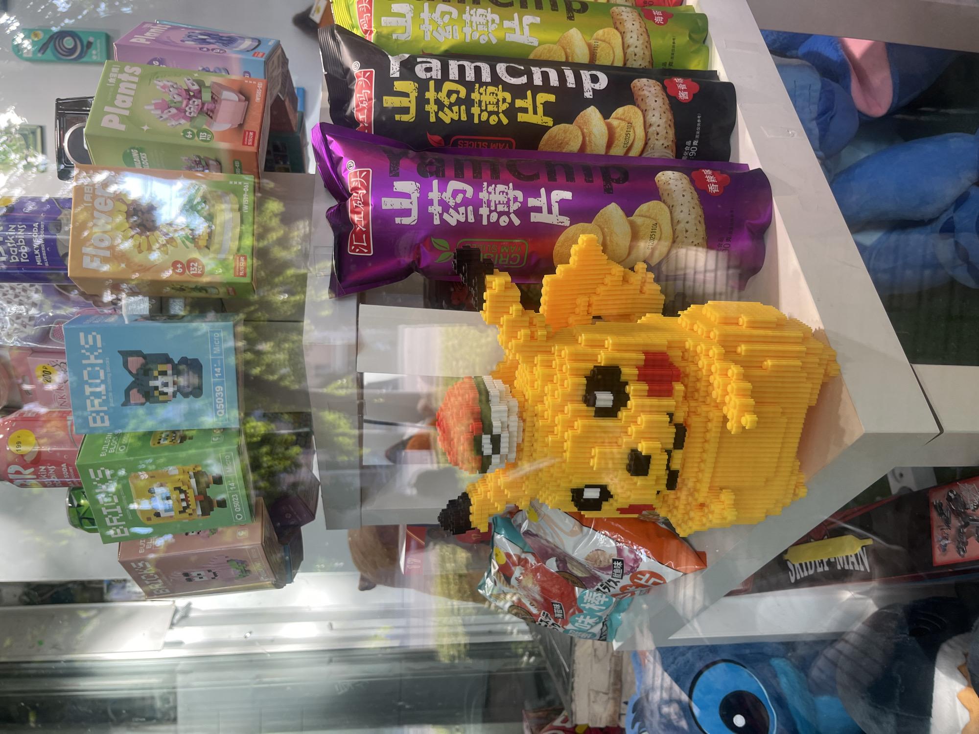 This image showcases a display of novelty building block sets and snack packages, likely in a store or vending machine. The building block sets feature miniature scenes and characters, with boxes labeled "Plants," "Flower," and "BRICKS." The snack packaging includes prominent text in what appears to be Chinese, with brands like "YAM SLICE" and "ramchip." One prominent object is a yellow, pixelated building block rendition of Pikachu, holding a red and white sphere resembling a piece of fruit or candy. The overall setting appears to be indoors, possibly a retail environment, with reflections on the glass surface indicating a bright, possibly daytime, environment. The items are arranged in a stacked, organized manner, suggesting a commercial display. There are no people visible in the image, and the focus is solely on the products.
