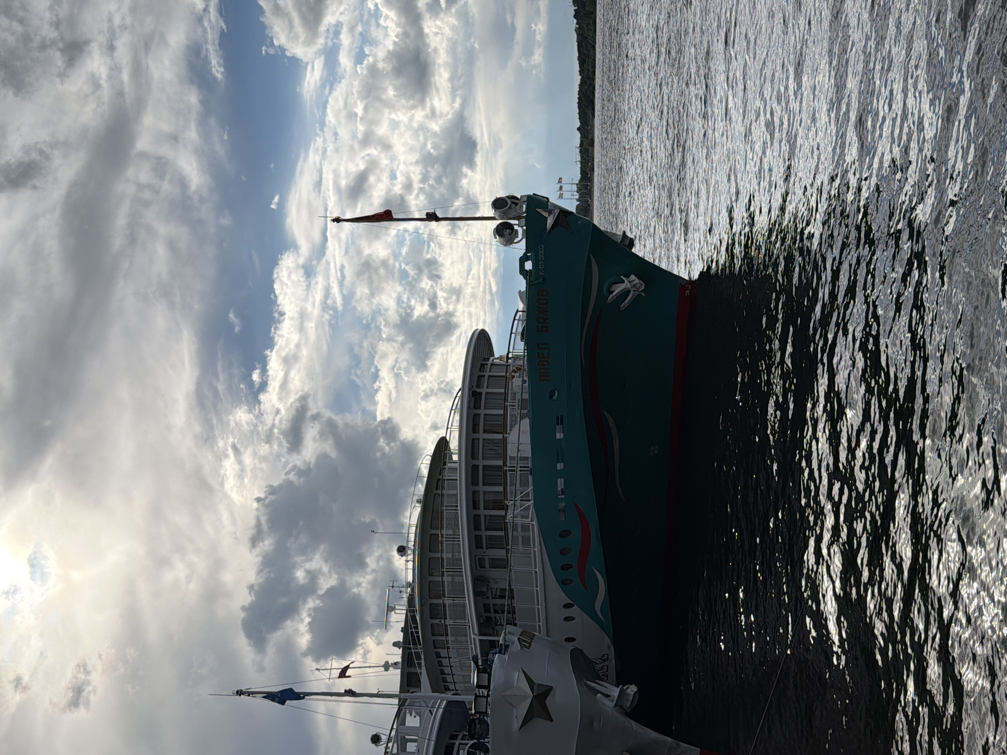 The image shows several boats docked on the water. The primary subject is a large, teal-colored passenger boat with white trim, featuring a red flag on its mast and a decorative bird emblem on its side. The boat has multiple decks with windows. To its left, another passenger boat, partially visible, is also docked. The scene is set on a body of water, likely a river, with a treeline visible in the distance on the right bank.
The sky is cloudy, with a mix of bright white and darker grey clouds, suggesting it might be late afternoon or early evening, with the sun partially obscured. The water is rippled and reflects the sky and the boats, creating a shimmering effect.
Visible text on the teal boat reads "ПАРЕЛ БАЖОВ" and "К-01-0002". The overall mood is peaceful and serene, characteristic of a quiet harbor setting. The location is identified as Perm, Russia. There are no people clearly visible in the image.