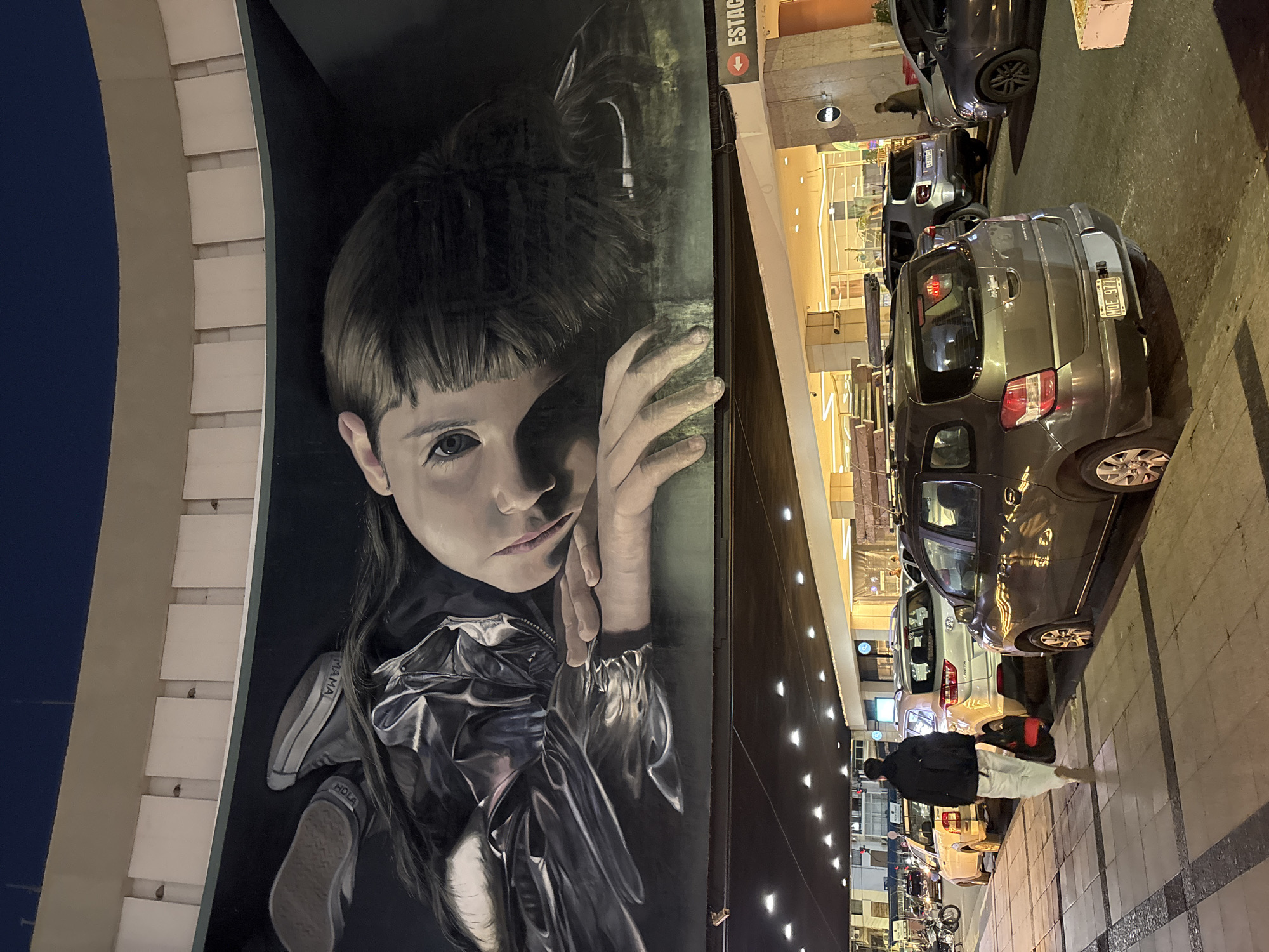This night-time image presents an urban scene, dominated by a large, realistic mural of a young girl painted on the side of a building. The mural depicts the girl with dark hair, prominent bangs, and large, expressive eyes, resting her chin on her hands. She wears a shiny, possibly metallic, silver jacket. Text is visible on her sneakers, reading "MAMA" on the side and "HOLA" on the sole (appearing upside down).
Adjacent to the mural is a paved sidewalk and a street, well-lit by artificial lights from buildings and street lamps, confirming the late hour. Several cars are parked along the curb. One silver car in the foreground displays a license plate with the registration "NOE 977," which is consistent with Argentinian vehicle plates. Further down the street, a person dressed in dark clothing is walking on the sidewalk, carrying a backpack. A red sign with a downward arrow and the partial text "ESTAC" (likely indicating "ESTACION," meaning station or parking) is affixed to a building overhead. The scene evokes a vibrant metropolitan environment, typical of Buenos Aires, Argentina, after dark.