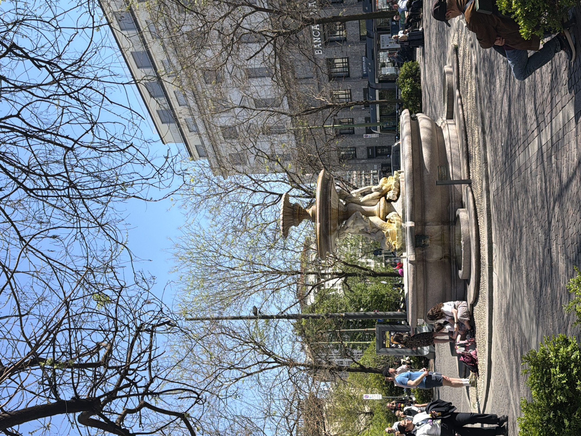 The image depicts a bustling scene near a fountain in Milan, Italy. The fountain, constructed from stone, has a tiered structure and figures sculpted near its top. People are visible near and around the fountain, some standing and interacting, while others are seated around the fountain. A paved walkway stretches out beside the fountain, with a building labeled "BANCA" in block letters towering in the background. The surrounding environment includes trees, some with bare branches and others with new green leaves. The sky is clear and blue. The photograph seems to have been taken on a sunny day.