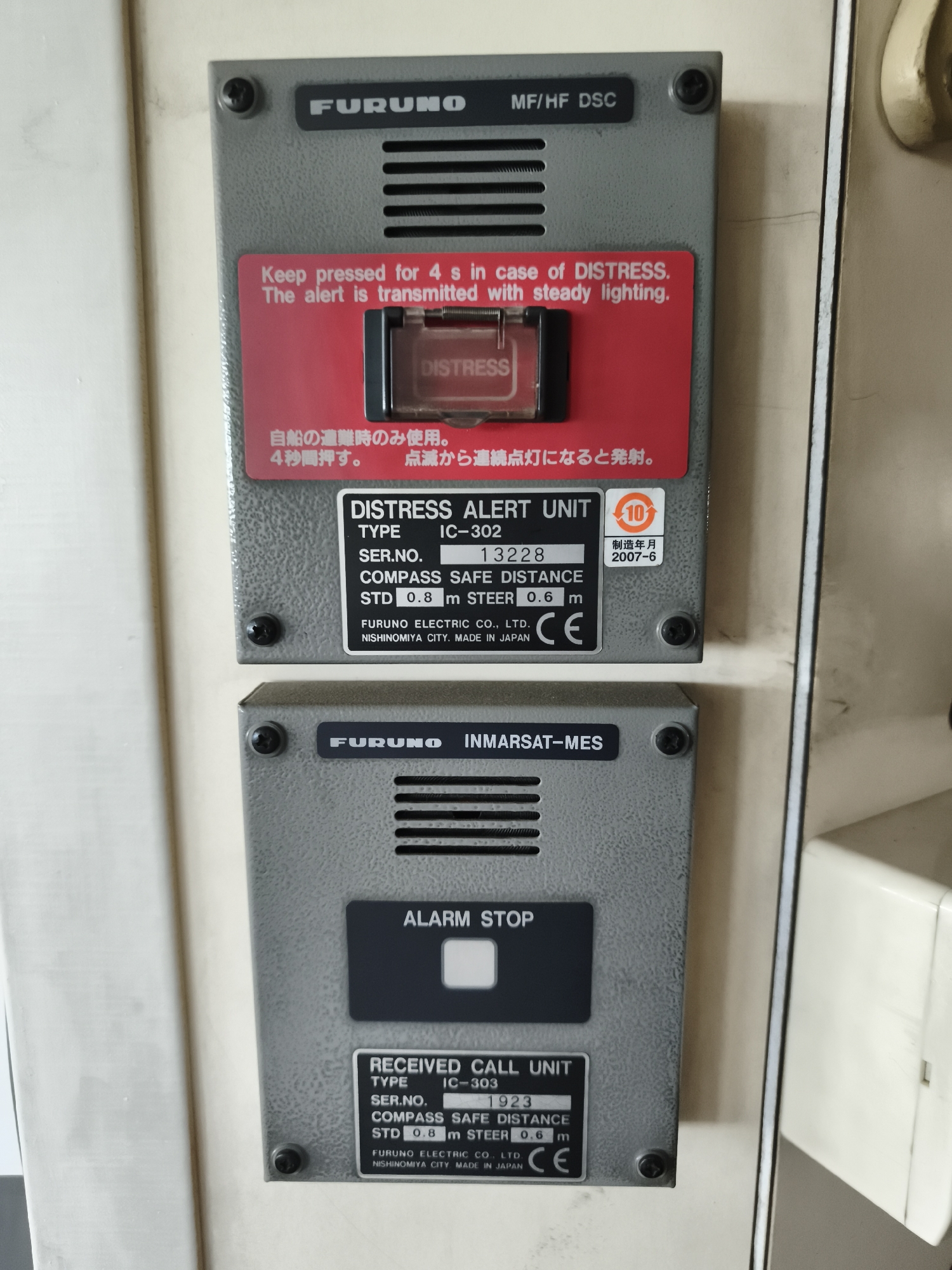 The image displays two Furuno electronic devices mounted on a beige wall. The upper device is a "DISTRESS ALERT UNIT" of type IC-302, with a serial number 13228 and manufactured in 2007-6. It features a red distress button with a clear protective cover. Below the distress button, there are instructions in English and Japanese. The lower device is a "RECEIVED CALL UNIT" of type IC-303, with a serial number 1923. It also has a label for "ALARM STOP". Both devices have "FURUNO" branding. The wall appears to be indoors, possibly in a control room or on a vessel. The image does not contain any people, and no specific location in Pirallahi Raion, Azerbaijan can be identified from the visual information.