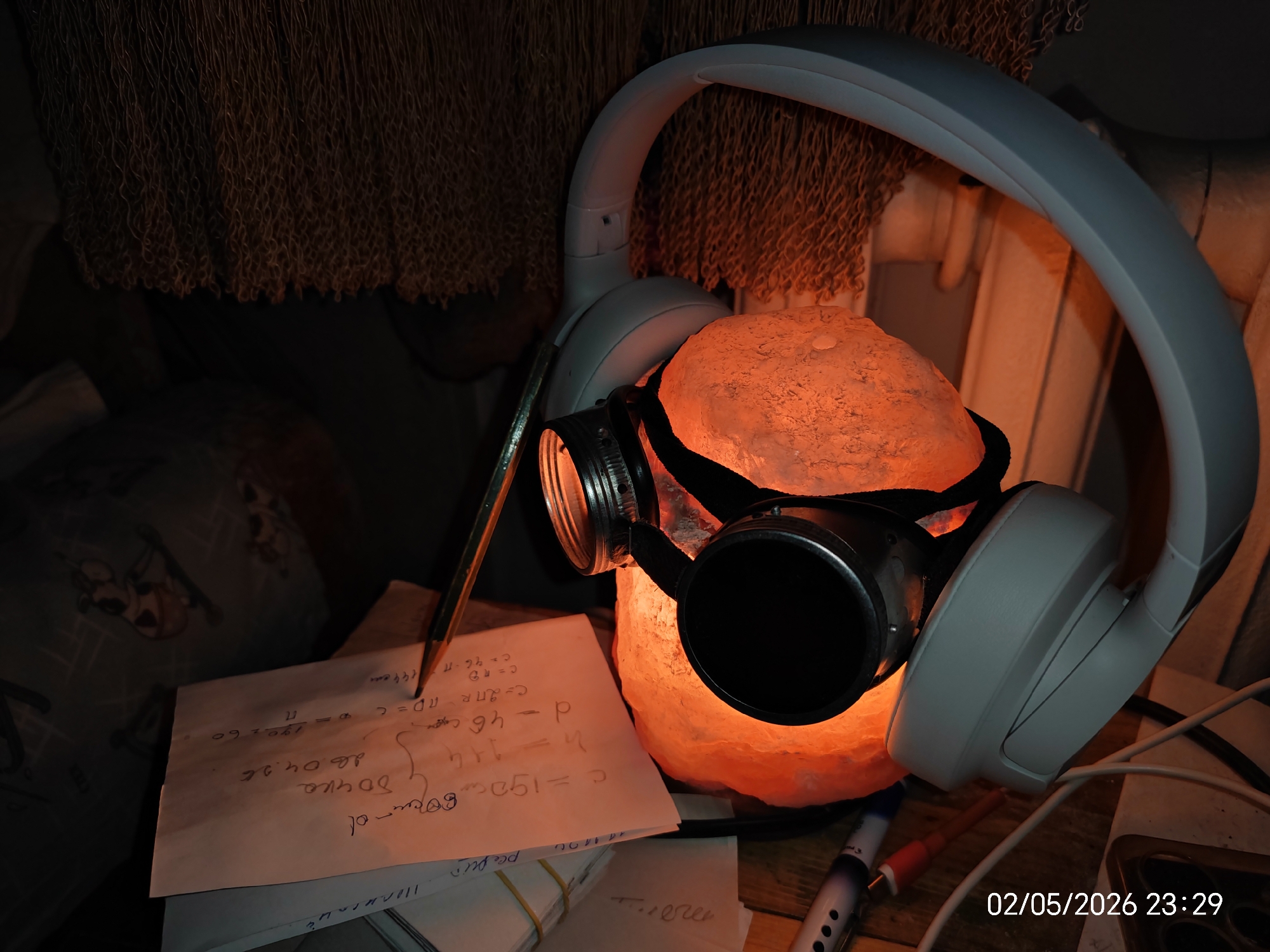 The image shows a pair of white headphones placed over an orange salt lamp which is wearing goggles. A sheet of paper with handwritten equations and notes is positioned to the left, with a pencil resting on it. Additional papers are stacked underneath. The date "02/05/2026 23:29" is visible in the lower right corner. The background is dark, with some textured objects visible.