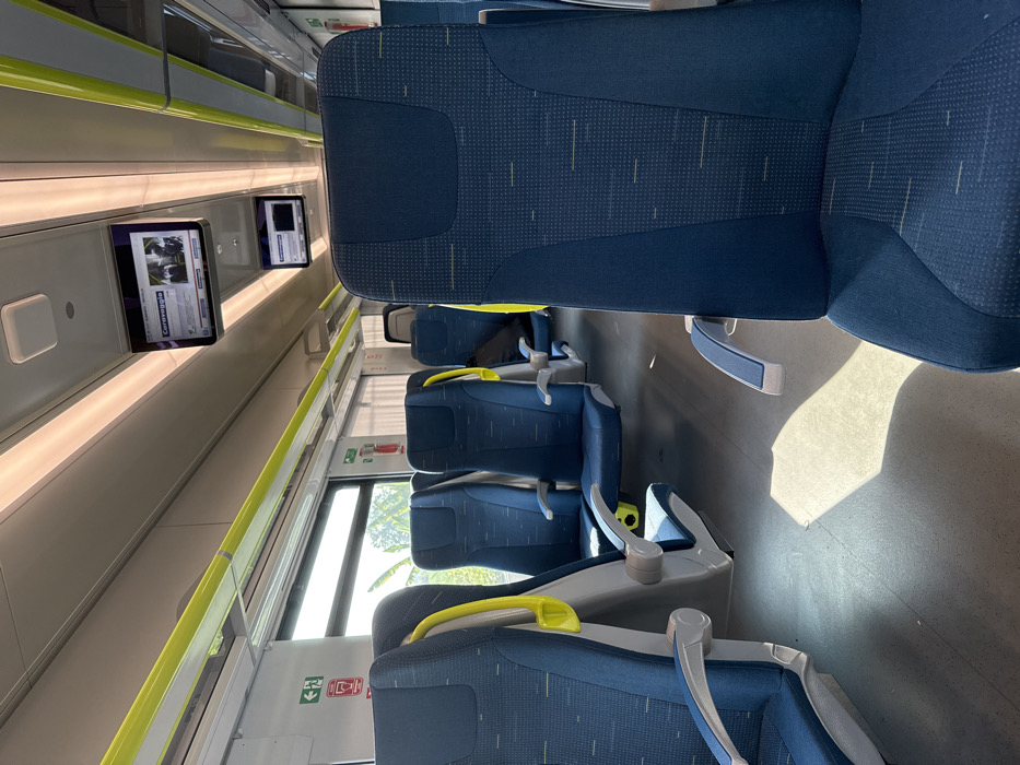 The image shows the interior of a train car in Caravaggio, Italy, located at coordinates 45.483 latitude and 9.646 longitude. The train car is empty except for a single passenger seat in the foreground, with a blue and grey patterned fabric cover. The seat has a grey armrest on the left side and a yellow armrest on the right. The train car is lined with a series of five other seats, all with blue and grey fabric covers. The train car is well lit by overhead fluorescent lights. There are three windows on the left side of the train car. The window furthest back in the image is partially obscured by a thin green curtain. The window in the center of the image shows a glimpse of trees and buildings outside. The window at the front of the image is obscured by a yellow handrail. There are two overhead monitors above the train seats; one monitor displays a news report and the other monitor is displaying a blank screen. The walls of the train car are painted white. The floor is covered in a light grey carpet. The train car is likely empty because it is not rush hour. The train car is clean and well-maintained. There are no visible signs of wear or tear. There are no personal items visible in the train car. The train car appears to be relatively new.