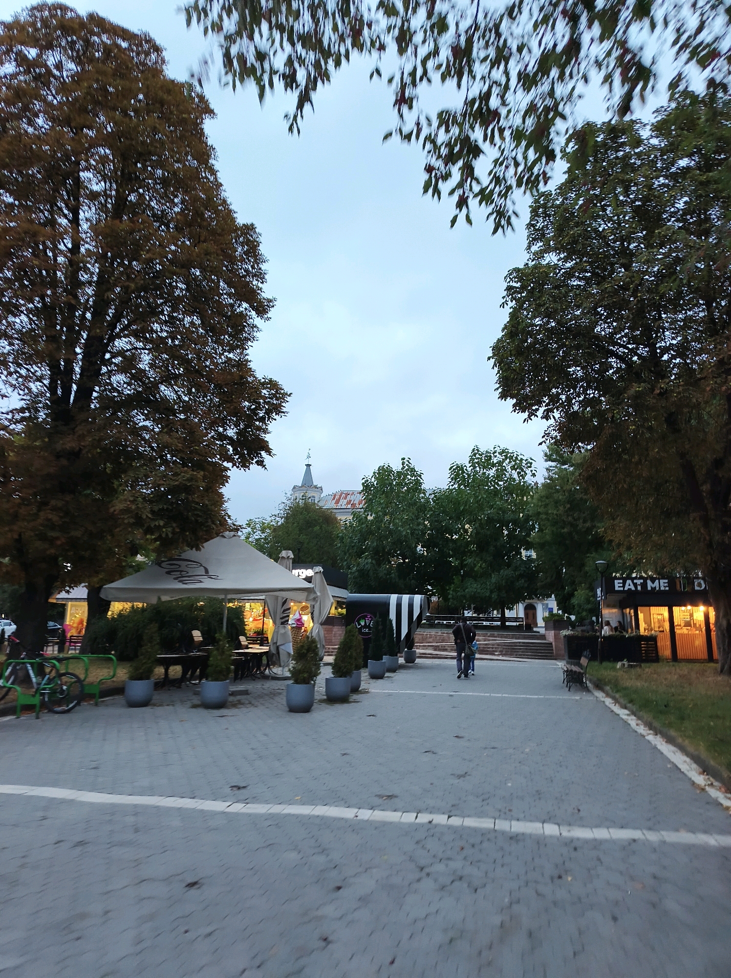 This is a vertical, medium-shot, eye-level photograph of a paved public walkway in Ternopil, Ukraine, during the day, likely early evening given the lighting. Two people, a man and a child, walk away from the camera towards a set of stairs. To the left, a large tree with brown leaves partially shades a white tent with tables and chairs set up underneath, possibly an outdoor cafe. To the right, a building with illuminated windows and a sign reading "EAT ME" is visible. The walkway is paved with interlocking grey stones and bordered by grass and trees. The sky is overcast.