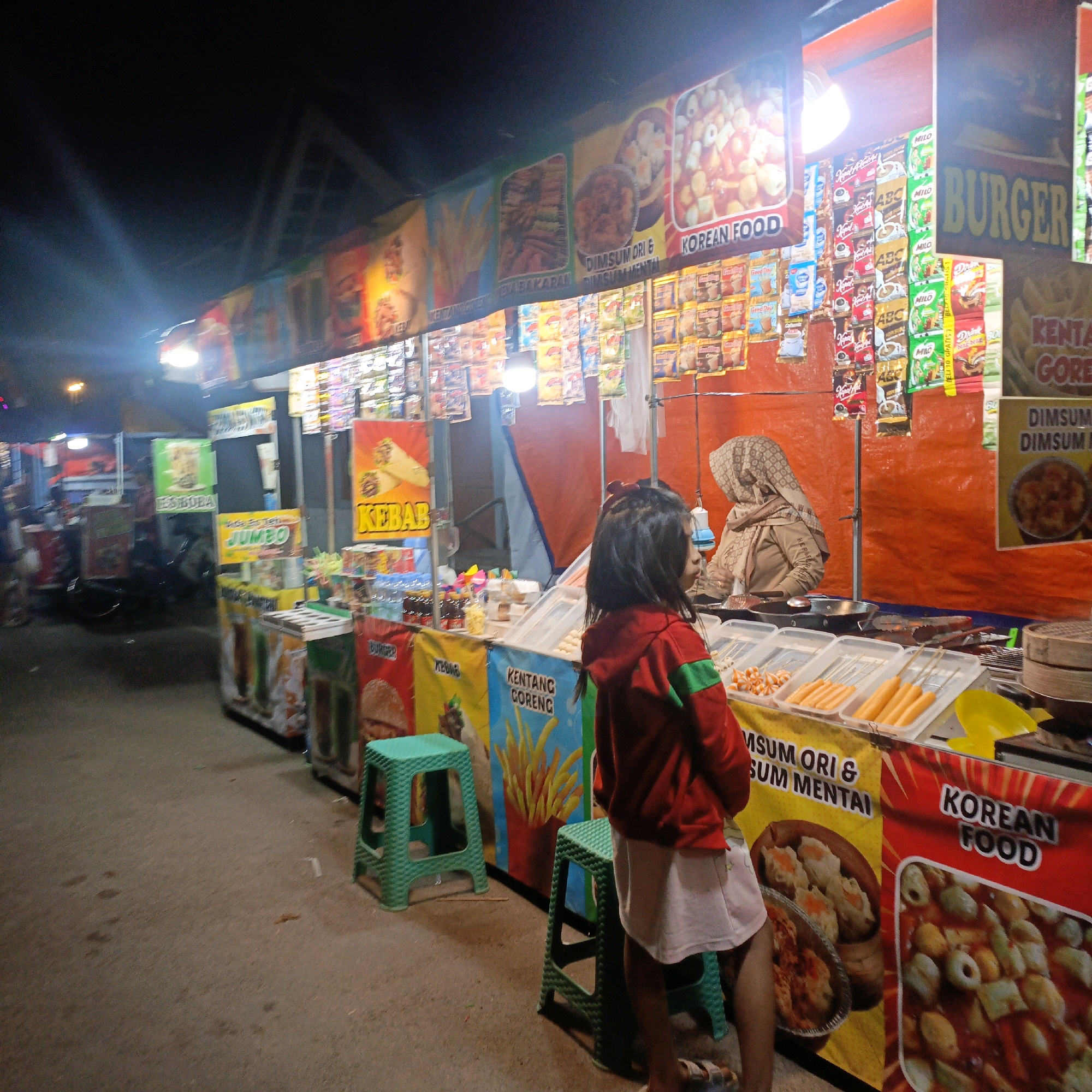 At night, in Salatiga, Indonesia, a food stall is set up outdoors. The stall displays banners advertising various food items, including "Kebab," "Jumbo," "Kentang Goreng," "Dimsum Ori & Dimsum Mentai," and "Korean Food," as well as "Burger." Various packaged food items are displayed on shelves above the stall counter. A young girl, wearing a red jacket and white skirt, stands near the stall, facing away from the camera. Behind her, a woman wearing a hijab appears to be attending the stall. In the foreground, green plastic stools are placed on the ground. The ground appears to be a paved outdoor surface. The lighting suggests it is nighttime, with artificial lights illuminating the stall.