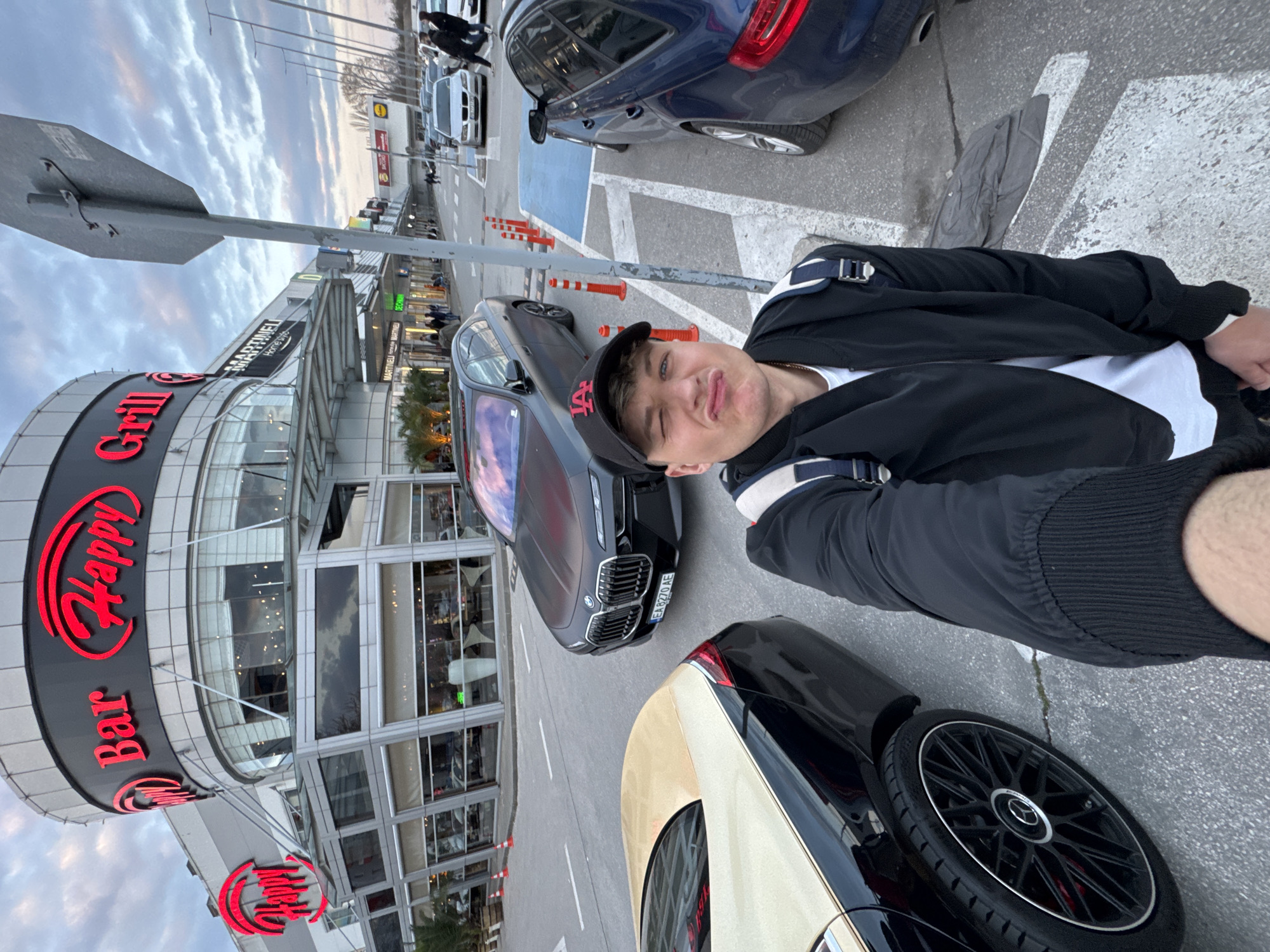 A male subject, approximately 20-30 years old, takes a low-angle selfie in an outdoor commercial area of Plovdiv, Bulgaria. He is positioned in the foreground, making a puckered facial expression with his left eye partially closed, wearing a black bomber jacket over a white t-shirt, a black baseball cap with a red "LA" logo, and a dark backpack.
To his immediate left, the rear quarter of a black convertible car with a gold-colored hardtop and black multi-spoke wheels featuring red brake calipers is visible. Behind him, a matte black BMW sedan (license plate "EA 8270 AE") is parked. To his right, the rear section of a dark blue vehicle with light-colored multi-spoke wheels is partially visible. The ground consists of asphalt and concrete with white and blue painted markings and several orange traffic cones. A gray metal signpost is prominently positioned to the subject's right.
The background features a modern, multi-story building displaying large red illuminated signs for "Happy Bar & Grill" and "MARTINELI Home style." Multiple individuals are seen walking in the distance near other parked vehicles. Further back, a "Lidl" supermarket sign is visible on an adjacent building. The sky is overcast.