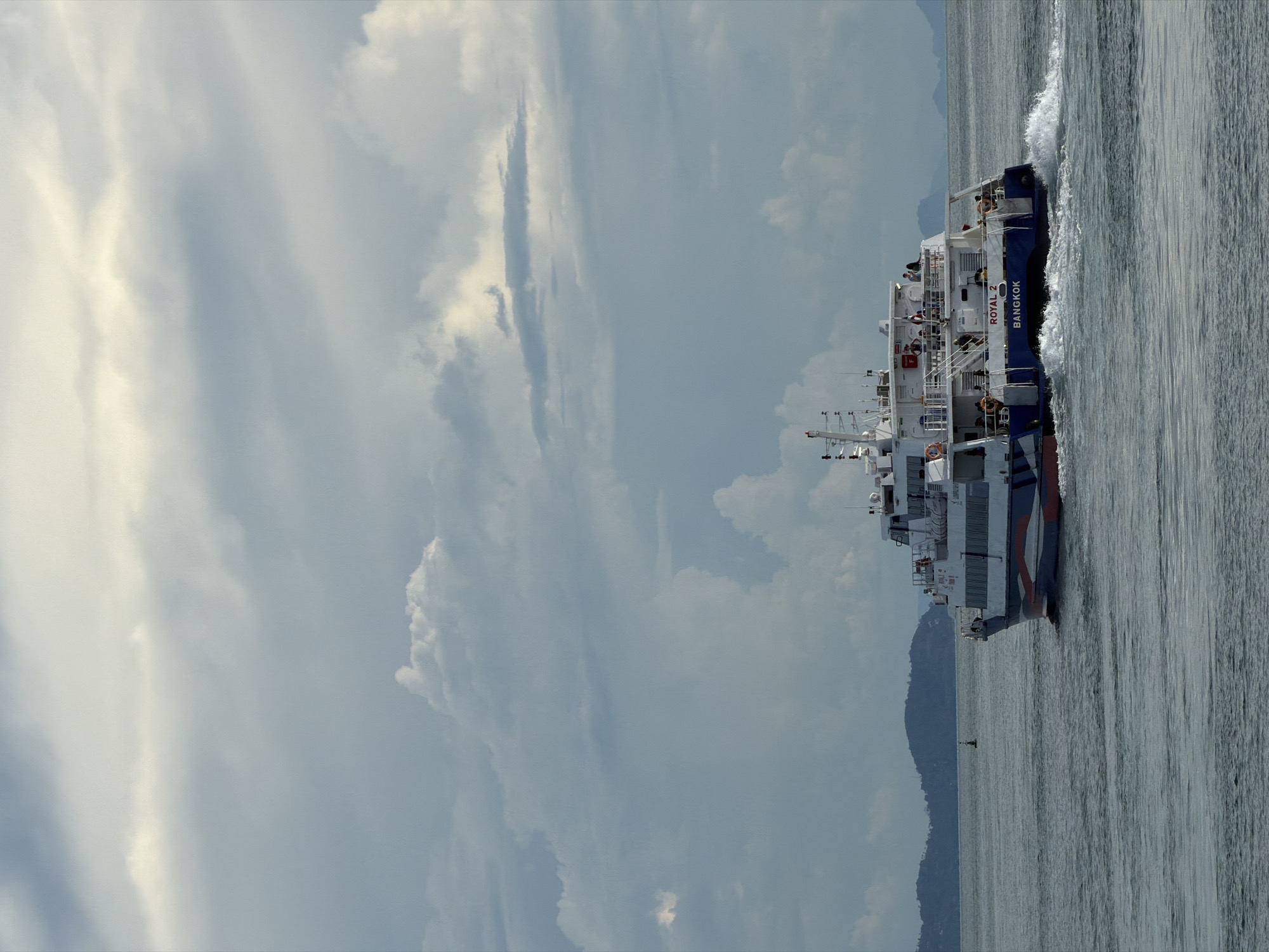 A white ferry, the "ROYAL 2 BANGKOK," travels across a body of water under a cloudy sky. The ferry, with blue and red trim along its hull, is mid-shot and moving from left to right. Several people are visible on the upper deck of the ferry, standing or walking. The water is rippled, with a white wake trailing the vessel. In the distance, a low-lying green hill or island is visible on the horizon, and a small green buoy floats in the water to the left of the ferry. The sky is filled with a mix of light and dark gray clouds. The scene appears to be set in a coastal area, possibly near Koh Phangan, Thailand, given the mention of Bangkok on the ferry and the tropical appearance of the distant landmass.