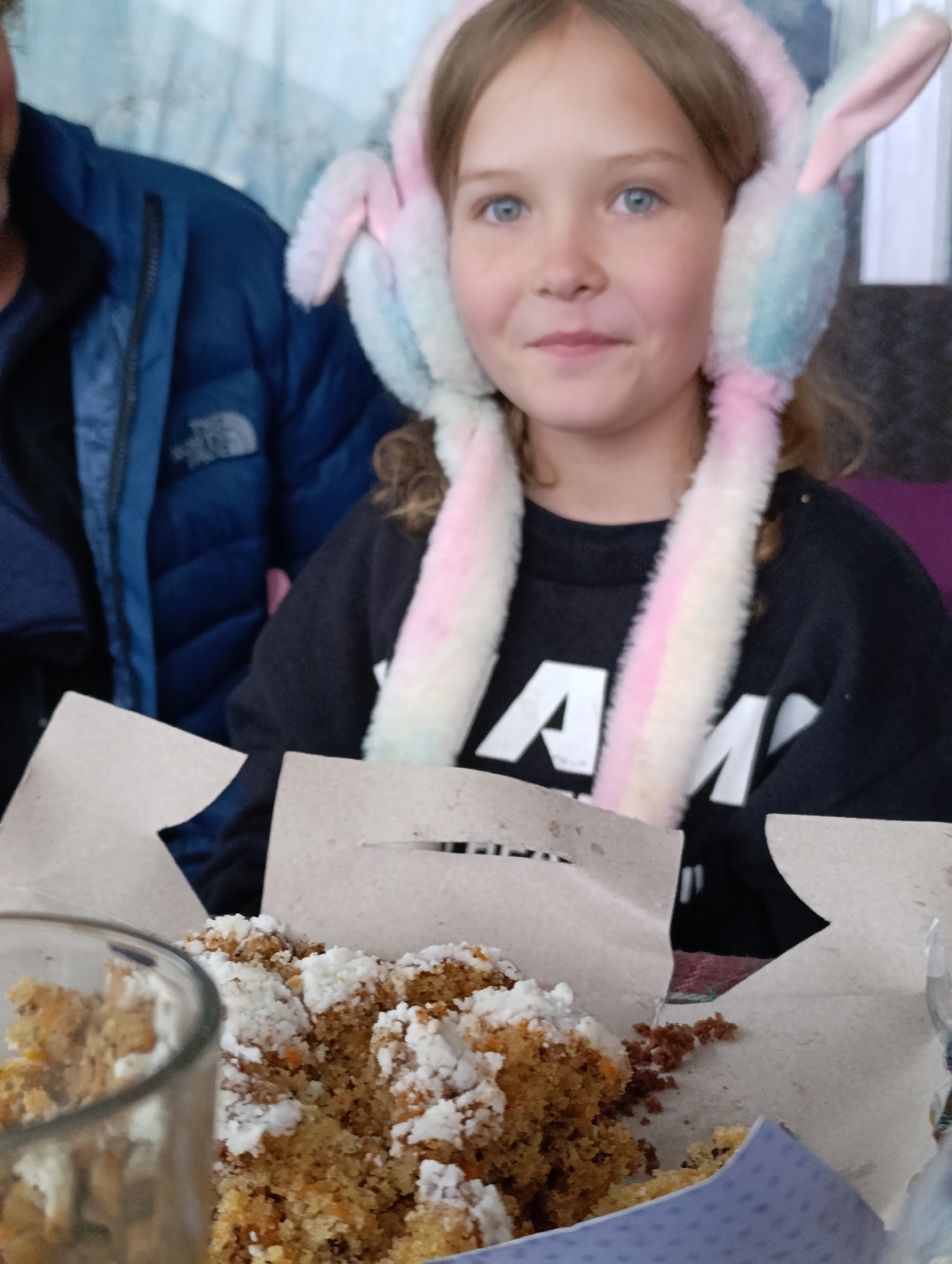In Vashisht, India, a young Caucasian girl with blonde hair and blue eyes is captured in a close-up shot, wearing fuzzy, pastel-colored earmuffs adorned with bunny ears. She is looking directly at the camera with a slight smile. In the foreground, a partially eaten cake, likely carrot cake due to visible orange flecks, is presented in a cardboard container, dusted with powdered sugar. A glass with some crumbs is partially visible on the left. Behind the girl, a person wearing a blue puffer jacket is visible on the left side of the frame. The background is blurred, suggesting an indoor setting.