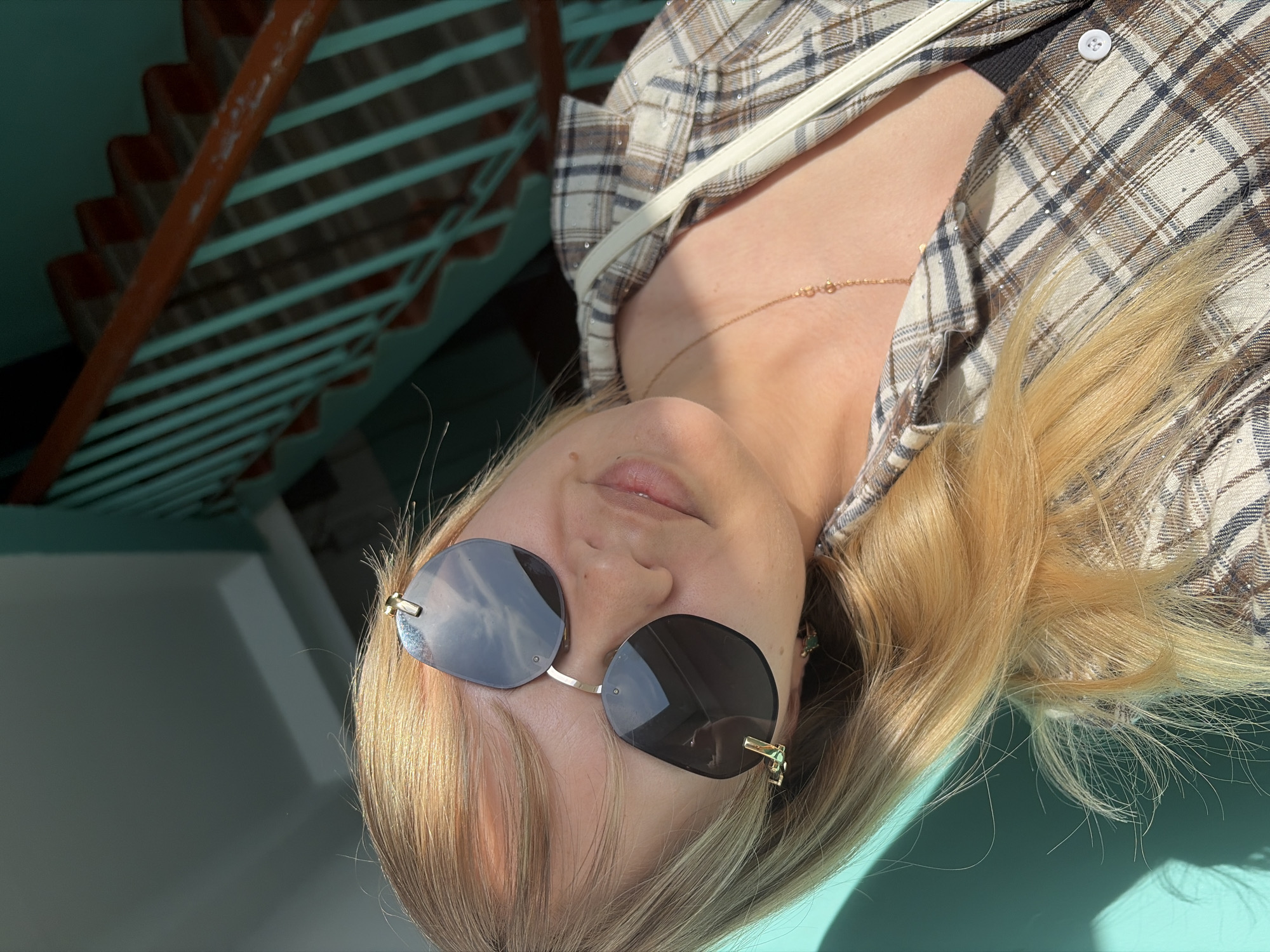 A woman with blonde hair, wearing sunglasses and a plaid shirt with a white strap across her shoulder, is the subject of the image. The background includes a blue-green wall and a staircase with orange handrails and blue-green steps. The location appears to be in the city of Chaikovsky, Russia.