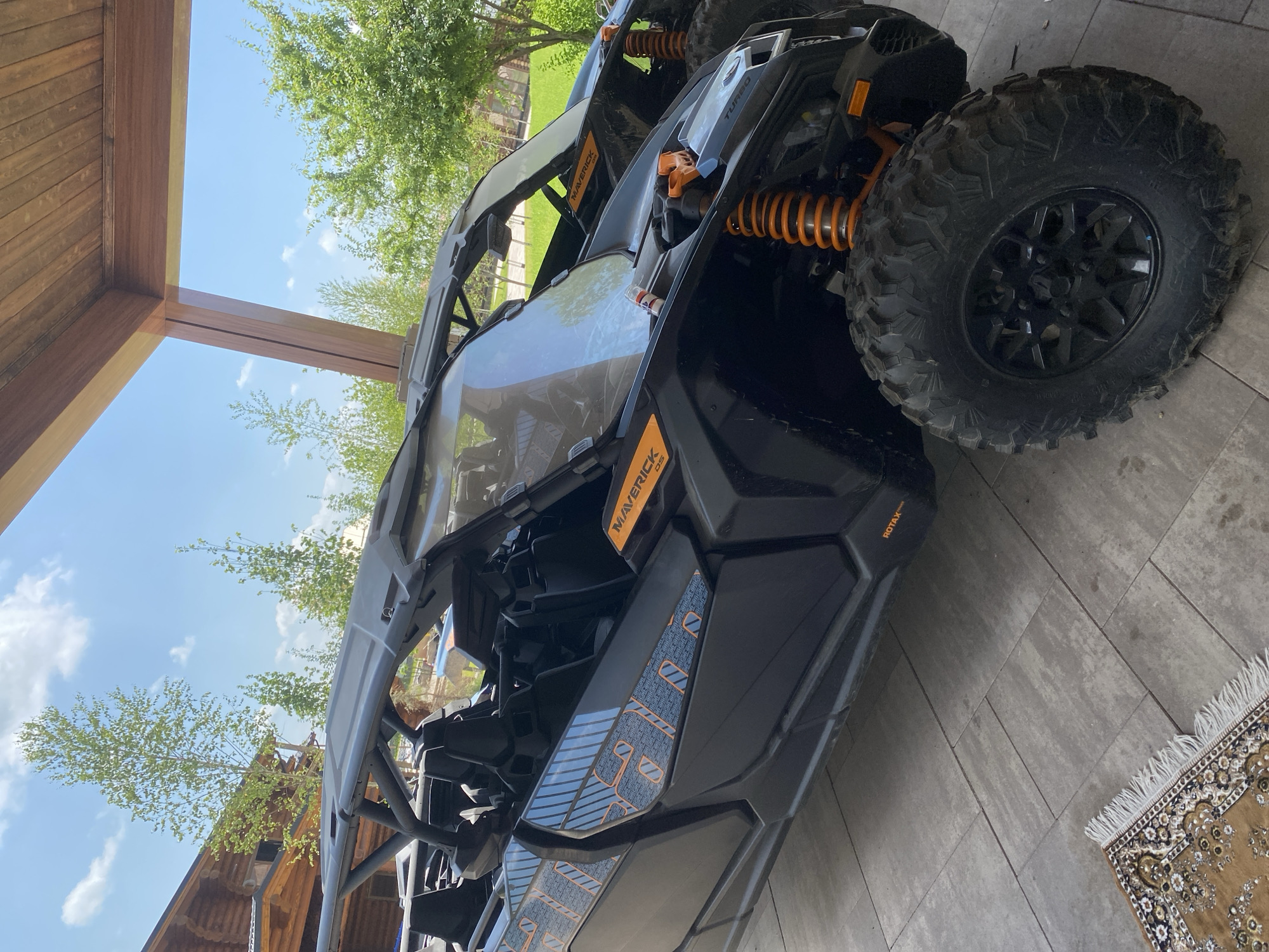 Two black Can-Am Maverick X3 side-by-side vehicles are parked under a wooden structure on a stone patio. The vehicles have orange accents on the suspension and trim. The one in the foreground has "MAVERICK" branding and a "ROTAX" logo. Trees and a log cabin are visible in the background. A small rug with a fringed edge is on the patio to the left of the vehicle. The sky is blue with scattered clouds.