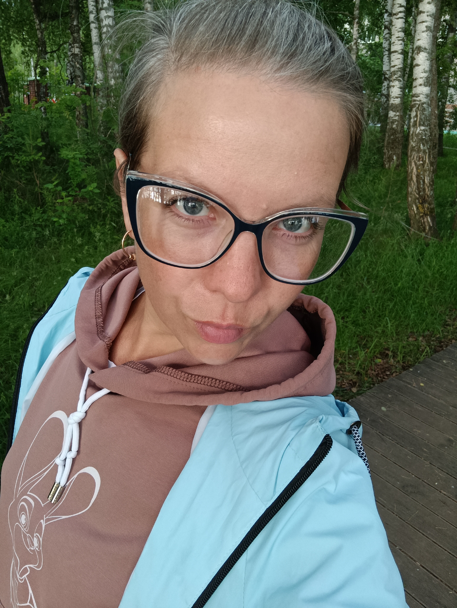 A woman wearing glasses and a light blue jacket over a brown hoodie with a cartoon rabbit print poses for a selfie outdoors. She has graying hair and is looking at the camera with a slightly pursed expression. The background consists of green grass, trees, and a wooden walkway, suggesting a park or outdoor recreational area. The image appears to have been taken in Chaikovsky, Russia, given the context provided.