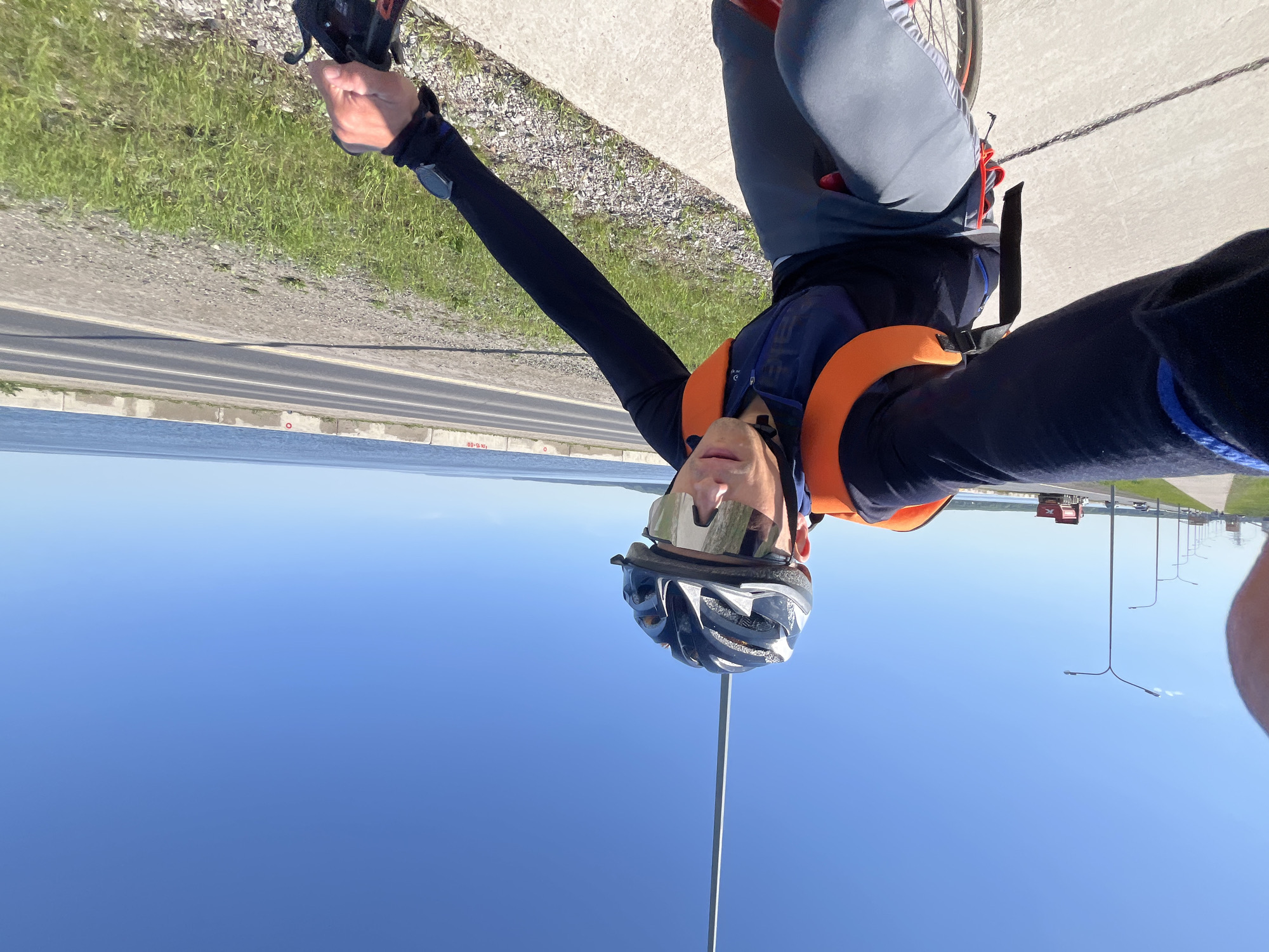 A cyclist wearing a helmet, reflective eyewear, a dark long-sleeved shirt, and an orange life vest is taking a selfie. The cyclist appears to be on a bridge or elevated road with a body of water to their left and a highway to their right, as evidenced by the presence of lampposts and passing vehicles. The setting is a sunny day with a clear blue sky. The location is Chaikovsky, Russia, suggested by the architecture and road markings.