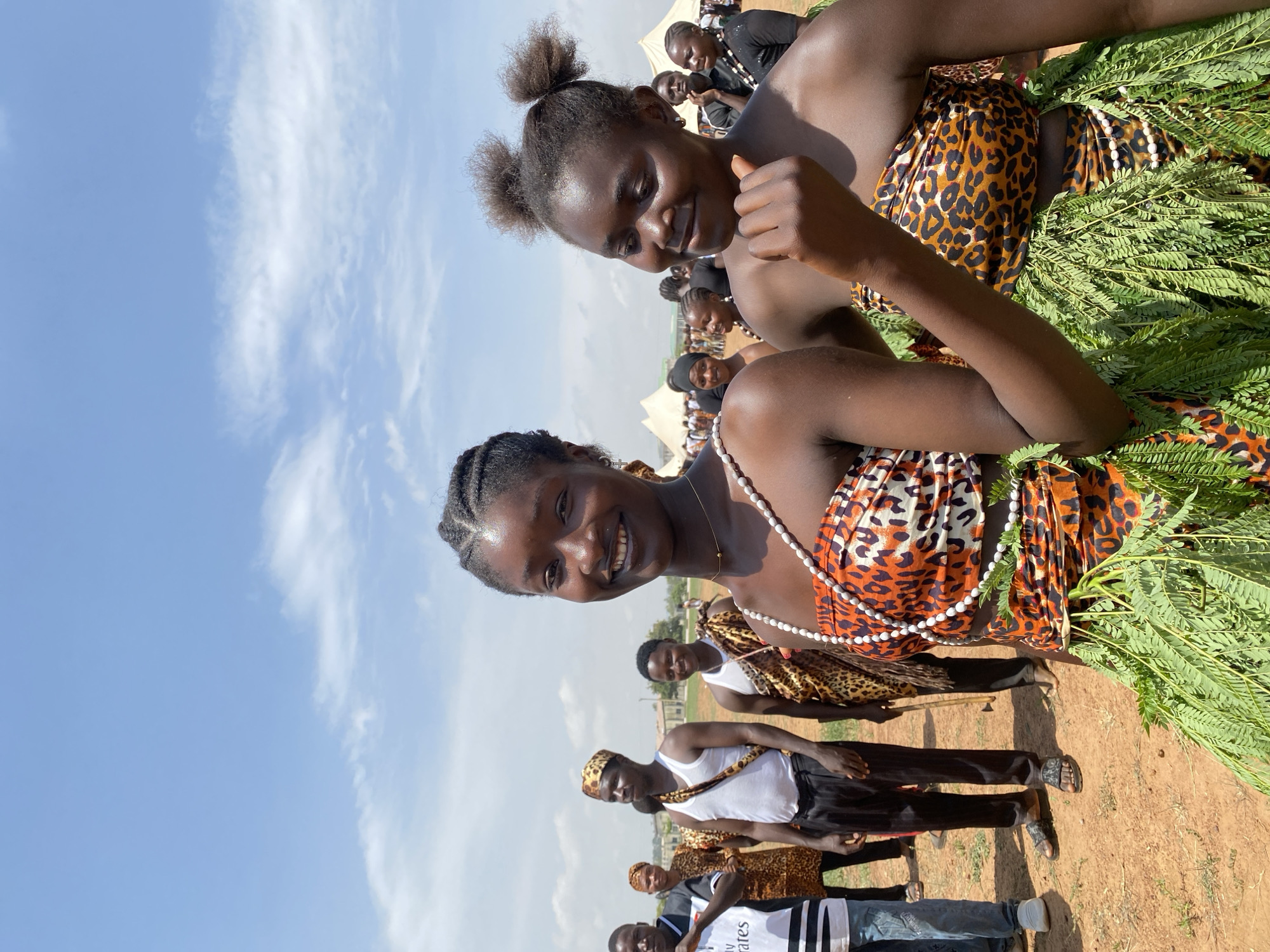 The image depicts a group of people outdoors under a sky with scattered clouds. Two women in the foreground are wearing leopard-print patterned dresses, one with her hair styled in two small buns and the other with braids. A necklace adorns the woman in front. Both are smiling at the camera. A bundle of green foliage is draped around the lower woman's waist. Behind them, other individuals are dressed in similar attire, some wearing white tank tops. A man in a white shirt with "Fly Emirates" printed on it is also visible. The ground appears to be dirt or reddish soil. It's not possible to verify the location as Durimi, Nigeria, from the image alone.