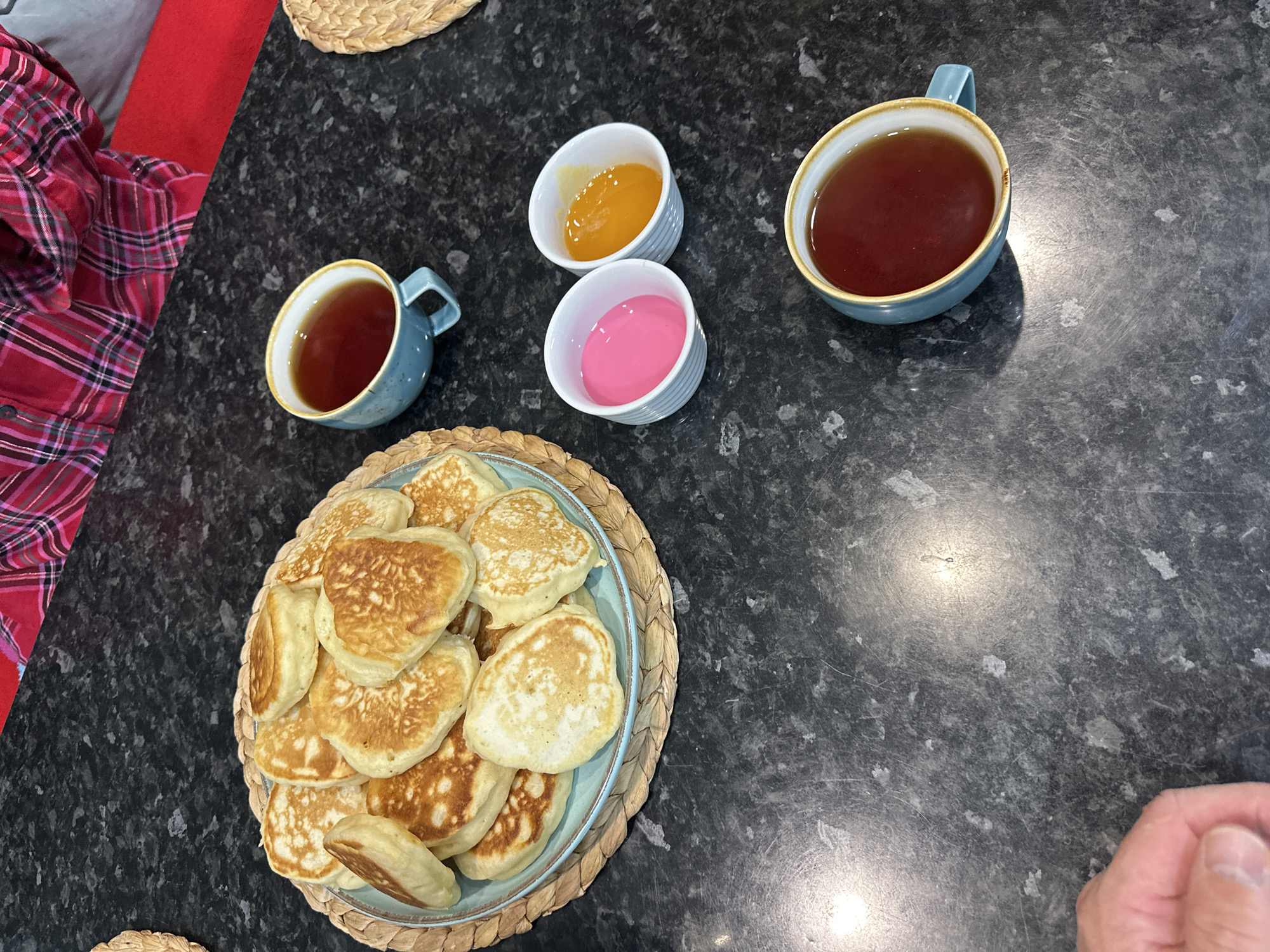 A top-down, medium shot captures a dark, speckled countertop with a meal laid out. In the upper left, a pile of golden-brown pancakes sits on a light blue plate, which rests on a woven placemat. To the right of the pancakes, two small white dishes contain brightly colored sauces, one pink and the other orange. Two blue teacups filled with dark liquid are positioned further right, one above the other. A person's hand, partially visible in the lower left, appears to be reaching towards the food. In the background, a person wearing a red and black plaid shirt is partially visible. The lighting suggests an indoor setting, likely a kitchen. There is no information to place this scene in Sochi, Russia.