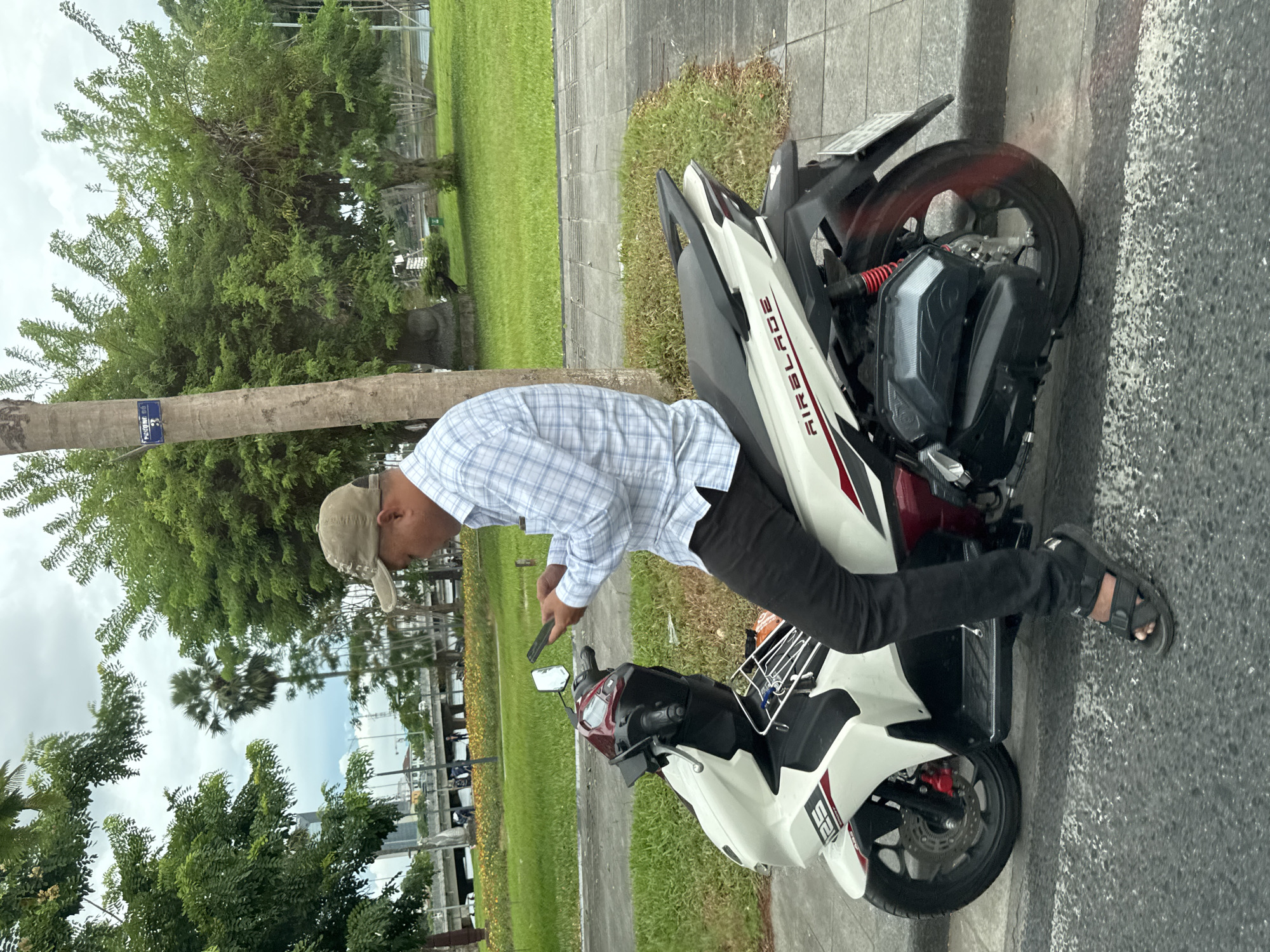 In Phú Xuân Ward, Vietnam, a man wearing a light blue plaid shirt, black pants, a beige baseball cap, and black sandals sits on a white and red scooter. He is looking down at his mobile phone, which he holds in his hands. The scooter is parked on a sidewalk next to a street. In the background, there are trees, a grassy area, a body of water, and a bridge with traffic. The sky is overcast.