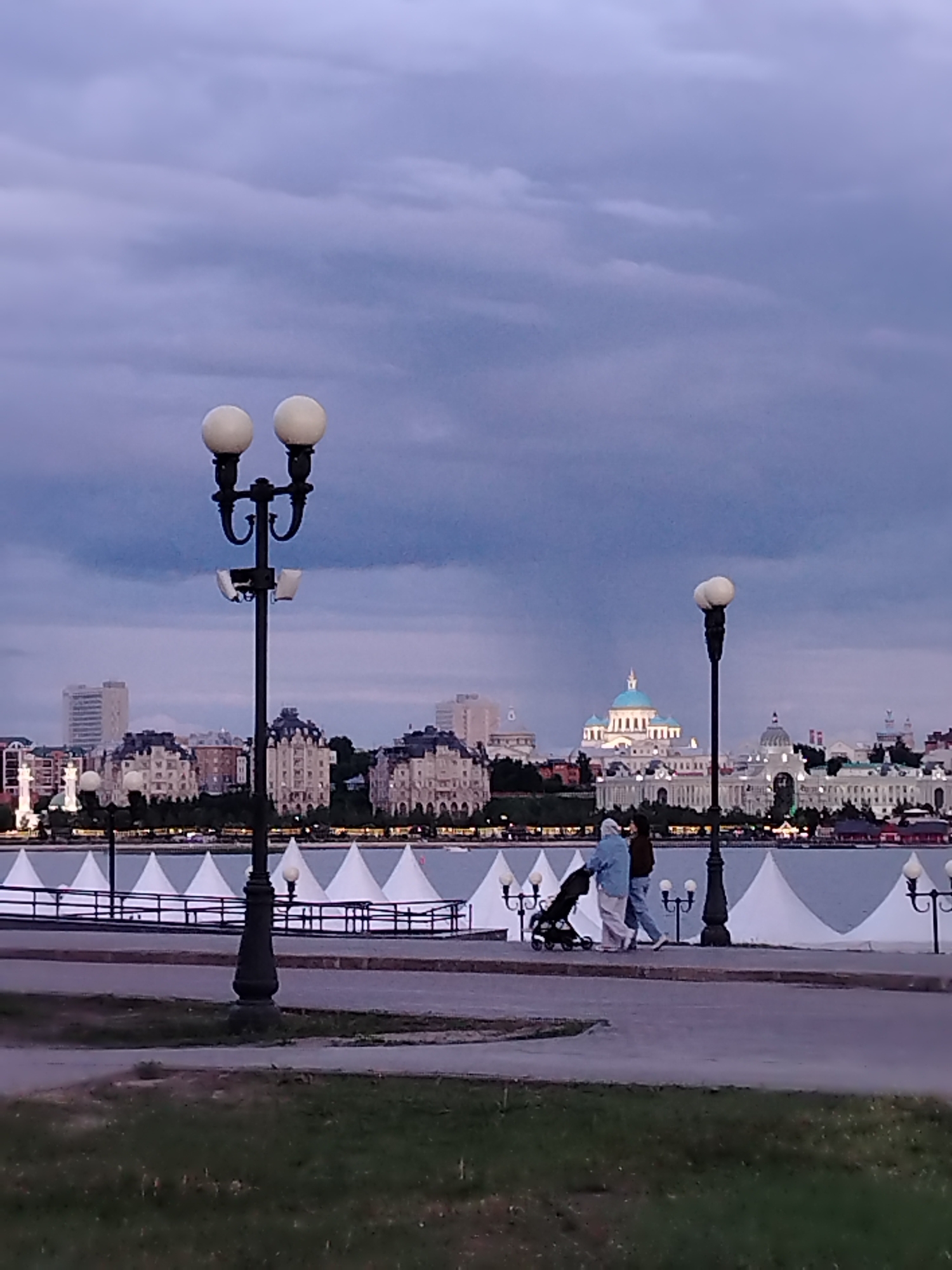 The image captures an evening scene along a waterfront promenade in Kazan, Russia, under an overcast sky filled with dark, purplish-gray clouds. The light suggests dusk, with artificial lights starting to illuminate the surroundings.
In the foreground, a paved path is visible, bordered by strips of green grass. Two prominent, traditional street lamps with multiple globe lights stand tall, one on the left and one on the right, providing illumination.
Across a body of water, the cityscape of Kazan stretches into the background. The most distinctive landmark is the Kazan Family Center, a large building featuring a light-blue dome and a classical white facade, centrally located in the distant skyline. Surrounding it are various buildings, some with traditional architecture, dark roofs, and intricate details, while others appear more modern. Along the water's edge on the far bank, a series of white, conical structures, possibly tents or event shelters, are arranged in a row.
In the mid-ground, two individuals are walking along the paved path. One person, dressed in a light blue top and light-colored pants, pushes a dark baby stroller. The other walks beside them, wearing a darker top and light pants. They appear to be taking a leisurely stroll.
There is no visible text in the image.