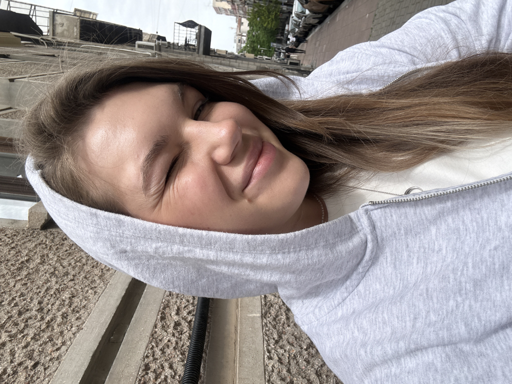 A young woman with brown hair, wearing a gray hooded sweatshirt, is smiling with her eyes closed. The location appears to be Belgrade, Serbia, based on the architecture of the building and street scene in the background. The woman is positioned on a textured surface, possibly a sidewalk or pavement. In the background, there are buildings, trees, and parked cars along a street. The sky is overcast.