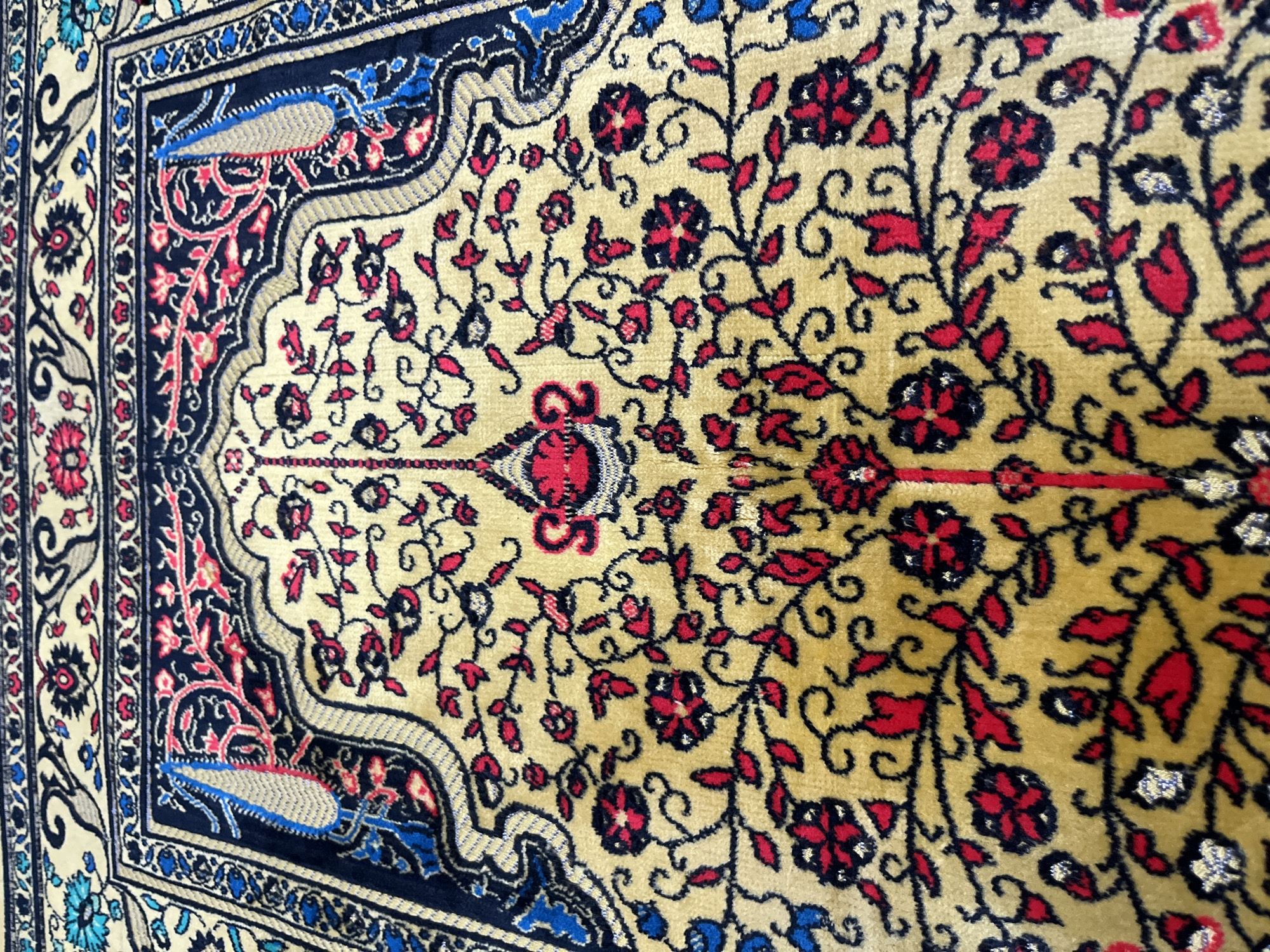 The image shows a section of a detailed oriental rug or carpet with a prominent design. The central field of the rug has a yellow/beige background adorned with floral and foliate patterns in red and black. A distinct curvilinear border with a dark blue background frames the central field. The border features stylized floral and leaf motifs in shades of red, blue, and white. The image is close-up, providing a detailed view of the rug's texture and design. No location information is included.