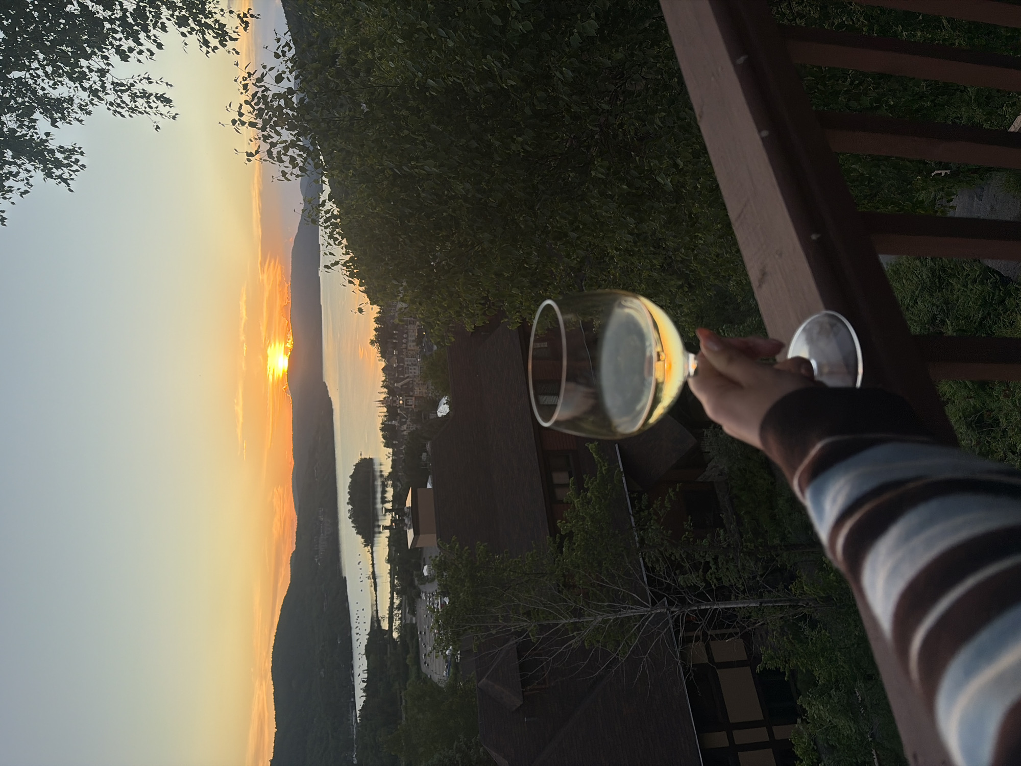 A first-person perspective shot captures a hand holding a wine glass filled with white wine, poised on a wooden railing. The glass reflects the vibrant orange and yellow hues of a sunset painting the sky. In the background, a picturesque panorama unfolds: a serene body of water, likely Lake Tremblant, is dotted with sailboats and a small island. Beyond the lake, the charming village of Mont-Tremblant, Quebec, Canada, is visible with its distinctive red-roofed buildings nestled among rolling green hills. Lush trees frame the scene, their leaves a deep green against the softening light of dusk.