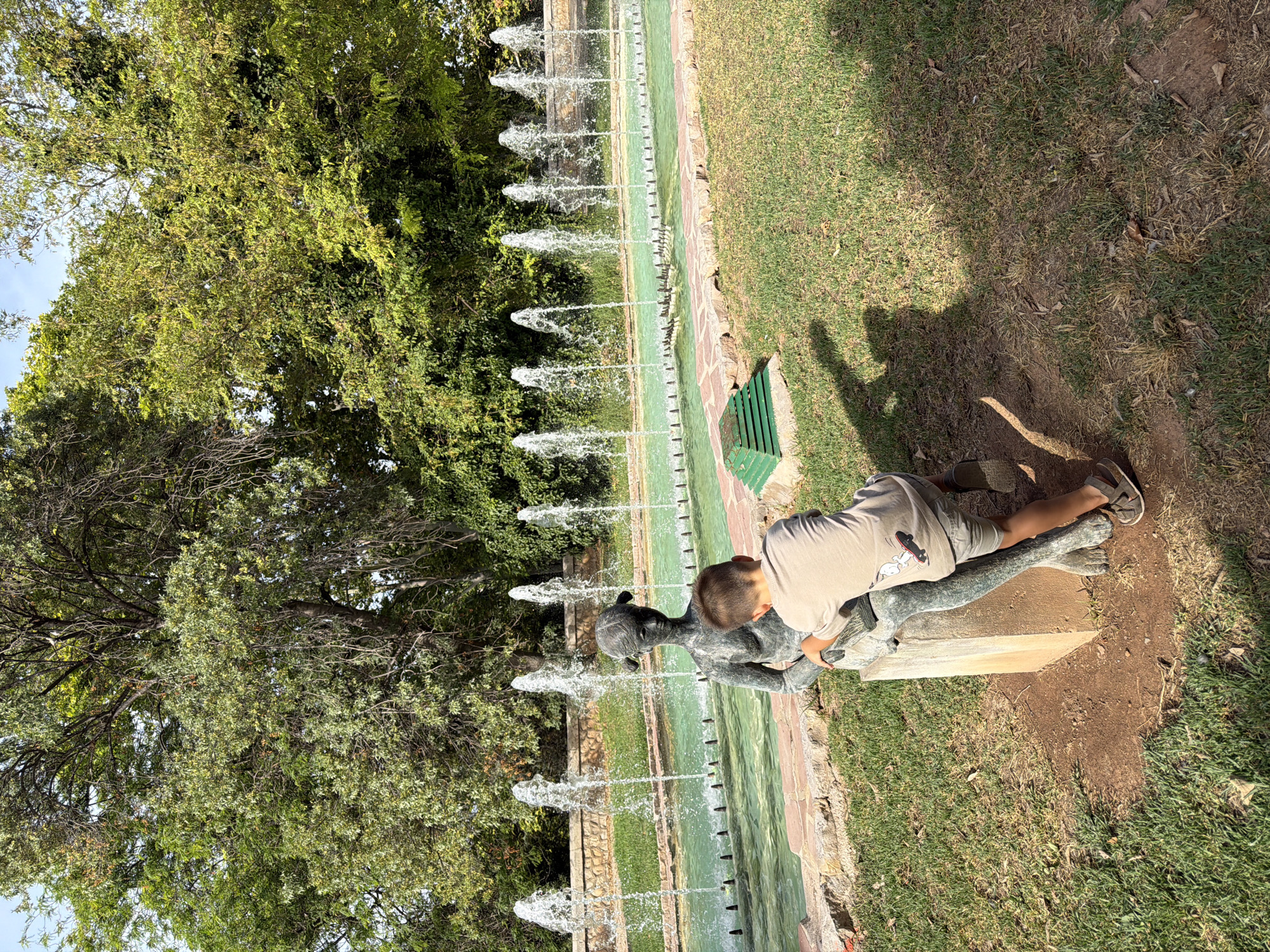 A young boy, wearing a light brown t-shirt with a cartoon graphic and gray shorts, leans against a bronze statue of a young woman. The statue is seated on a concrete pedestal. The boy's left arm is extended, and he appears to be touching the statue. In the background, a long, rectangular fountain with multiple jets of water creates a visual barrier. Beyond the fountain, a stone wall and lush green trees form the backdrop. The ground is covered in grass and patches of dirt. The scene is illuminated by bright sunlight, casting shadows on the grass. The location is outdoors in a park-like setting.