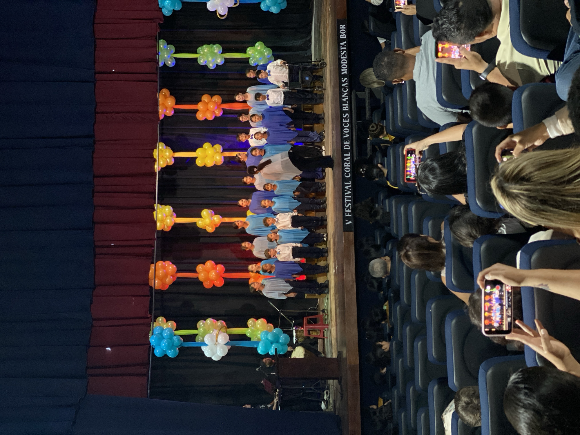 This image captures an indoor choral performance, likely an evening event, held in an auditorium or theater in Juan Griego, Venezuela. The scene centers on a stage, which is decorated with colorful balloon arrangements resembling flowers in shades of blue, white, green, orange, and yellow. These lighted balloon structures are vertically aligned along the stage's edge, set against a dark curtain backdrop.
On the wooden stage, a group of approximately twenty children and young adults are visible, dressed in various light blue, white, and darker blue tops, suggesting a choir uniform. They stand in rows, facing the audience, poised for or engaged in a performance. One child on the stage is seated in a wheelchair. Towards the lower left of the stage, musical instruments and equipment, possibly a keyboard stand and other audio gear, are set up, with a person seated nearby, likely providing musical accompaniment.
A prominent vertical banner along the right side of the stage displays the text: "V FESTIVAL CORAL DE VOCES BLANCAS MODESTA BOR." This identifies the event as the "5th Choral Festival of White Voices Modesta Bor," highlighting a cultural or educational gathering focused on young vocalists.
The audience occupies rows of dark blue upholstered seats in the foreground and mid-ground. Numerous spectators are actively holding up smartphones, recording or photographing the performance on stage, indicating a high level of engagement, pride, and enthusiasm for the event. The atmosphere appears supportive and celebratory.