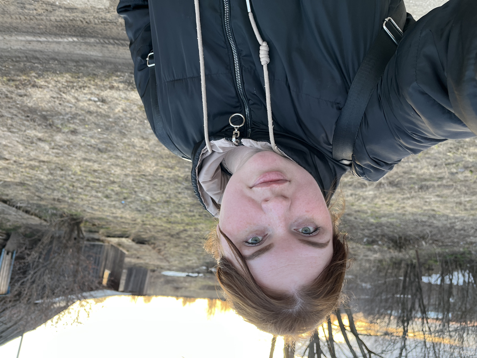 A woman is taking a selfie outdoors in what appears to be a rural or semi-rural area. The woman is wearing a dark, quilted jacket with a light-colored inner layer and a shoulder strap across her body. She has light skin, light eyes, and reddish-brown hair. The background consists of bare trees, a wooden structure that resembles a shed or small building, and what looks like a field or unpaved ground. The light in the image suggests it might be sunrise or sunset. Based on the prompt, the location is Павловка, Russia.