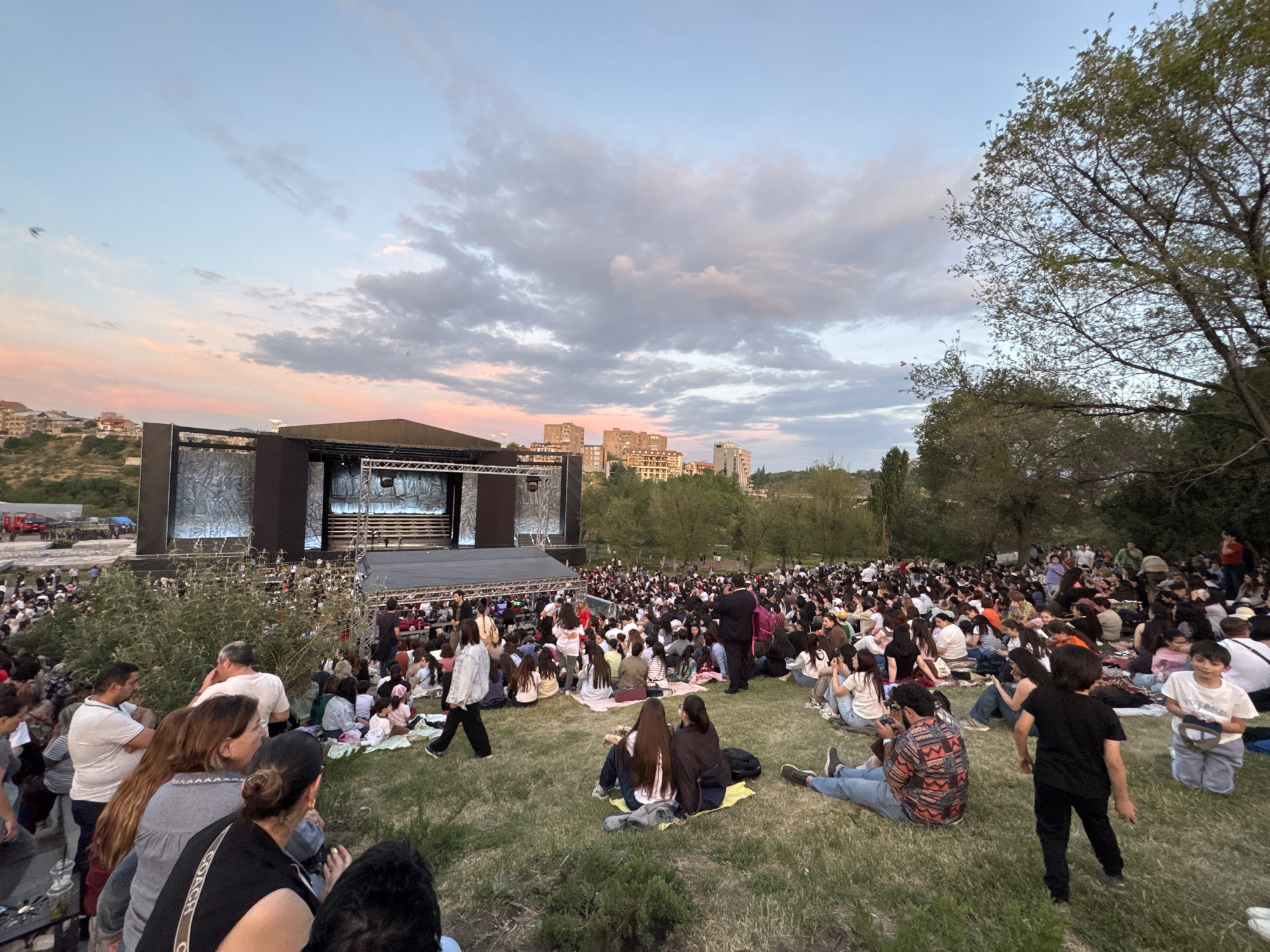 This image depicts a large outdoor gathering in a park during twilight hours, likely an evening concert or performance, located in the city of Yerevan, Armenia. A substantial, dark-colored stage structure is prominently positioned on the left side of the frame, featuring multiple levels, visible lighting rigs, and what appear to be screen panels, with performers present on the main platform. A dense crowd, estimated to be several hundred to over a thousand individuals, occupies the grassy slope in front of the stage. Most attendees are seated directly on the grass, some on blankets or mats, and are oriented towards the stage. The audience consists of a mixed demographic, including adults, teenagers, and some children; some individuals are standing or walking through the crowd. In the foreground, several individuals are clearly visible, seated and standing on the grass. In the midground, the main body of the audience extends across the park. The background reveals hills dotted with numerous multi-story residential buildings characteristic of urban landscapes, along with a significant cluster of trees on the right side of the frame. The sky exhibits a gradient of light blue, soft pink, and gray clouds, indicating the late afternoon or early evening.
