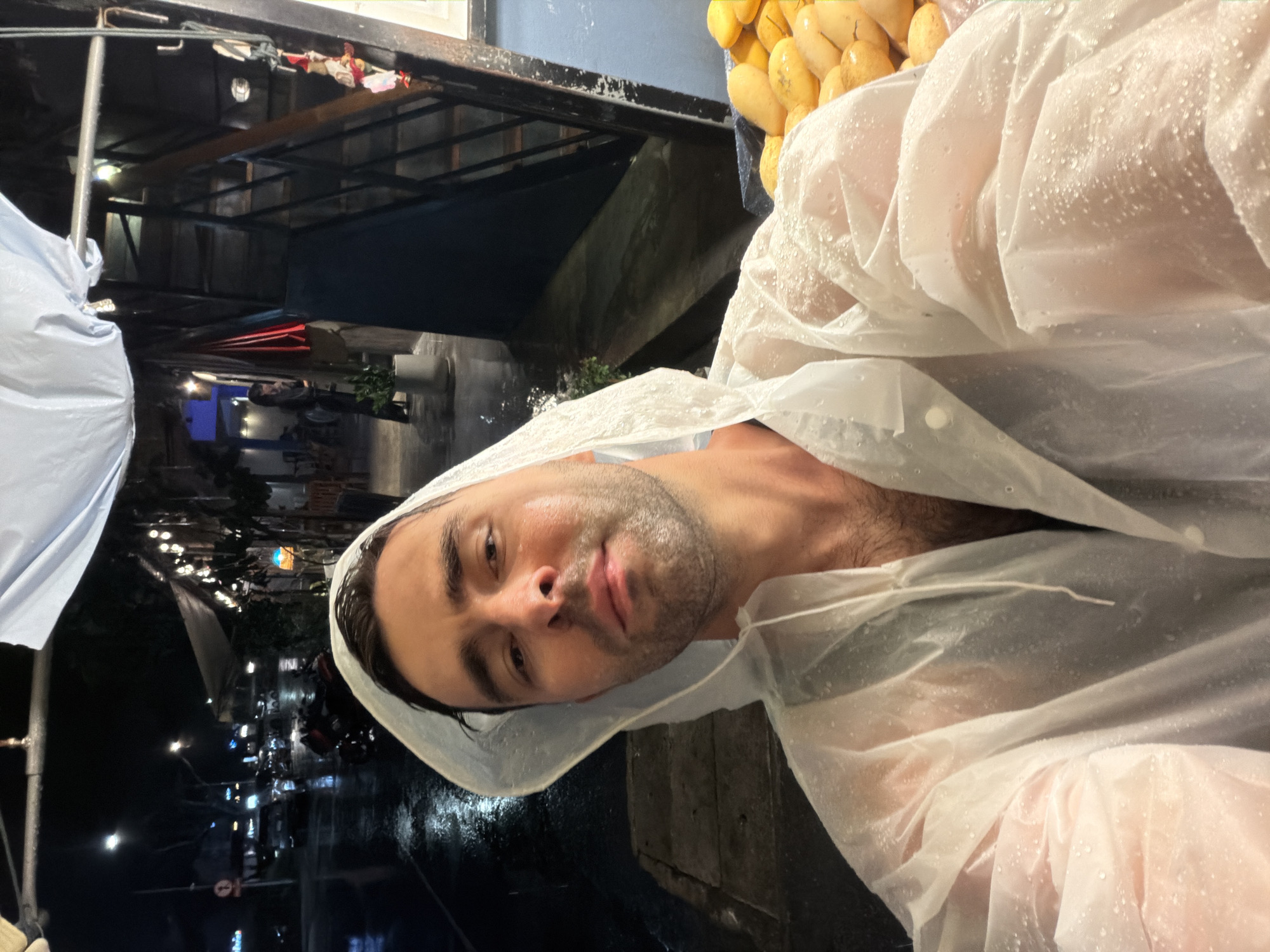 A man wearing a translucent white hooded raincoat poses in what appears to be the city of Choeng Thale, Thailand, based on the street scene in the background. The street is illuminated with lights, and there are vehicles and pedestrians visible. A display of yellow fruits, possibly mangoes, is located to the upper-right of the man. Water droplets are visible on the raincoat.