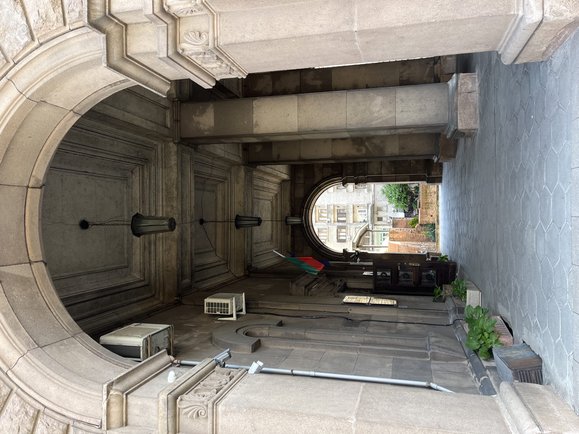 The image shows an exterior view of a structure in Sofia, Bulgaria. The structure is constructed of gray stone, likely limestone or granite, featuring prominent architectural details such as arches, beams, and moldings. The scene is framed by a large archway on the left, which gives way to a covered area leading to an opening in the distance.
Inside the covered space, there are various objects, including what appear to be air conditioning units, a black cabinet, and a flag with colors red, green, and white, resembling the Bulgarian flag. The space appears to be a type of portico or entry point into a building or courtyard.
The background visible through the distant opening reveals buildings and greenery, suggesting an urban environment. The overall impression is that of an aged and somewhat neglected urban space, showcasing classic stone architecture with signs of modern installations.