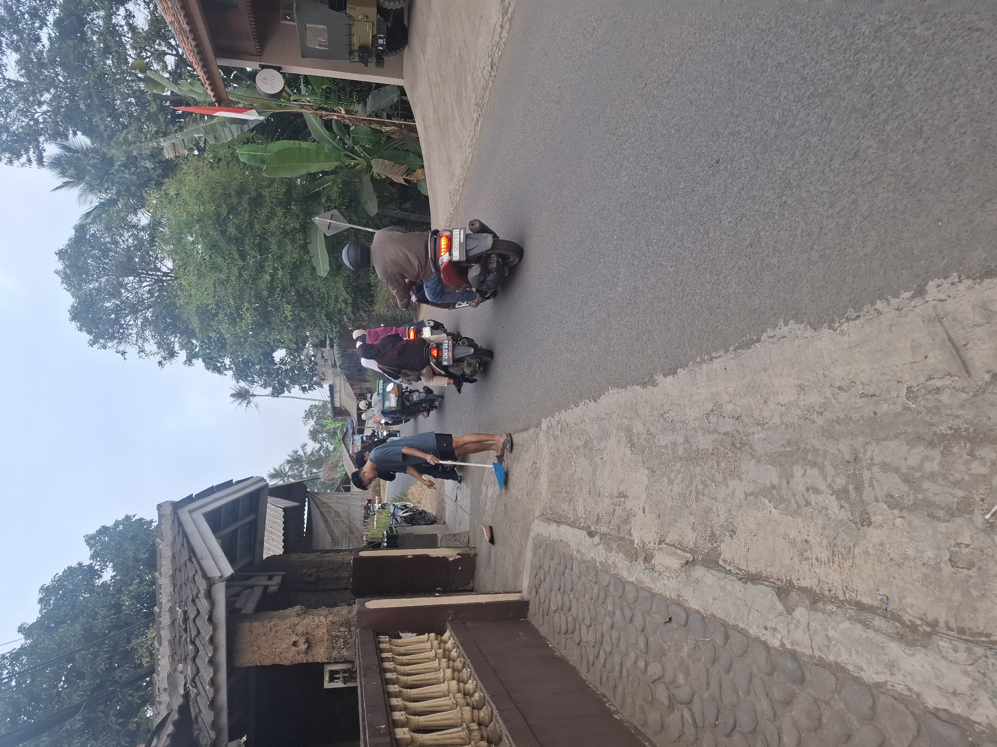 The image depicts a street scene in the village of Ciheulang Tonggoh, Indonesia, likely during the daytime. A young man, wearing a blue t-shirt and dark shorts, is sweeping the concrete sidewalk with a blue dustpan and broom. To his left, a section of a building with a tiled roof and stone-like facade is visible, with a decorative balustrade running along the edge of the sidewalk. In the background, several motorbikes are traveling along the asphalt road. Some of the riders are wearing helmets and carrying passengers, with one motorbike having a delivery box on the back. Behind the road, lush green trees and vegetation are abundant, characteristic of a rural or semi-rural Indonesian environment. A building with a red tile roof and an Indonesian flag is also visible on the right side of the road, along with what appears to be a parked military vehicle. The sky is overcast, suggesting a potentially humid or cloudy day.