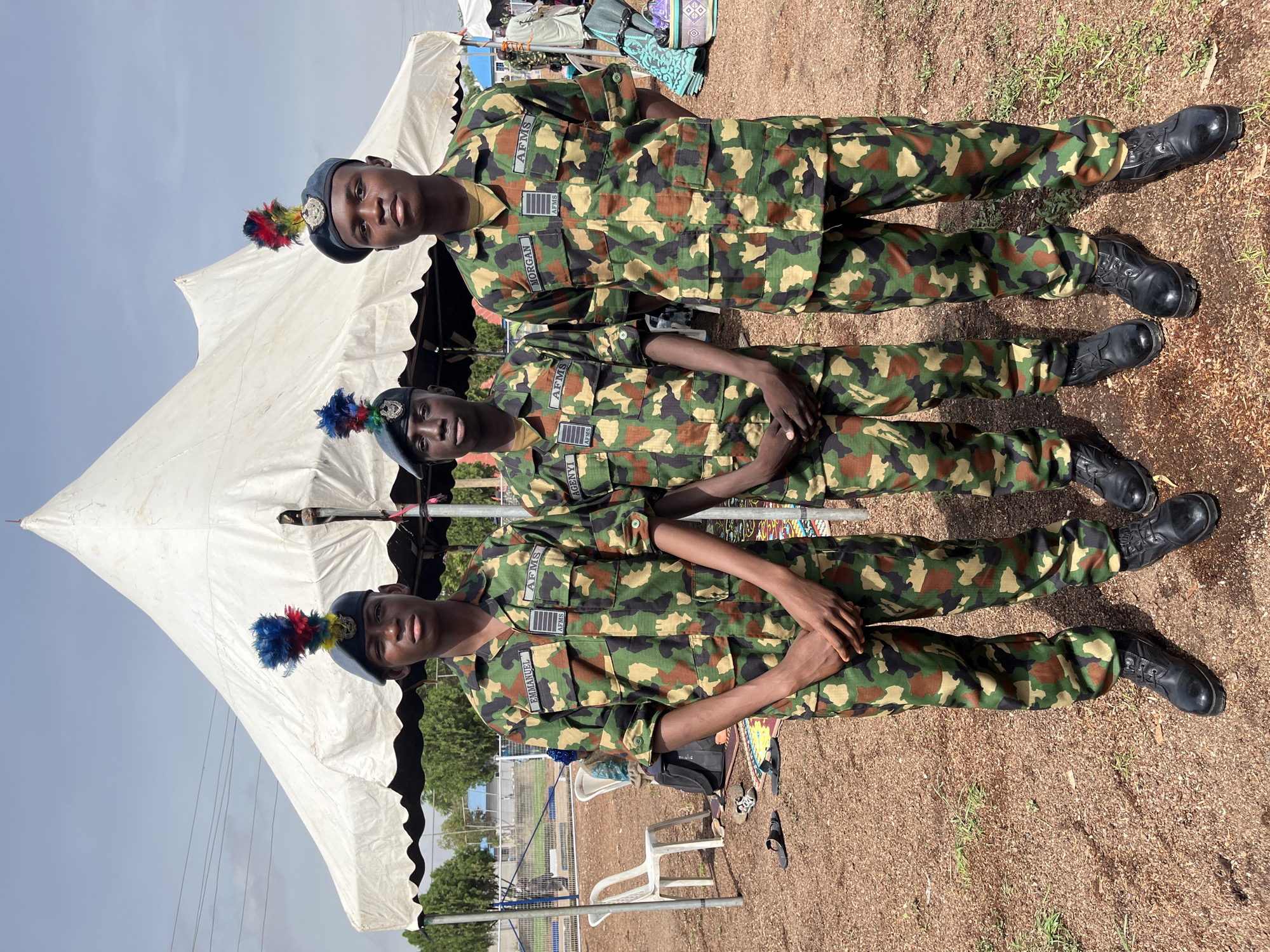 Three young men in camouflage military uniforms stand in front of a large white tent under a cloudy sky. Each man wears a dark beret with a colorful feather plume. The man on the left has a name tag reading "EMMANUEL" and another tag with "AFMS". The middle man's name tag is "MUHAMMED" and also has "AFMS". The man on the right has a name tag reading "MORGAN" and "AFMS". All three men are looking towards the camera. They are standing on a ground covered with reddish-brown dirt and small stones. To the left of the men, there are white plastic chairs and bags. In the background, there are trees and some structures. The overall scene suggests an outdoor event or ceremony. The location is Barakin Lerawi, Nigeria.
