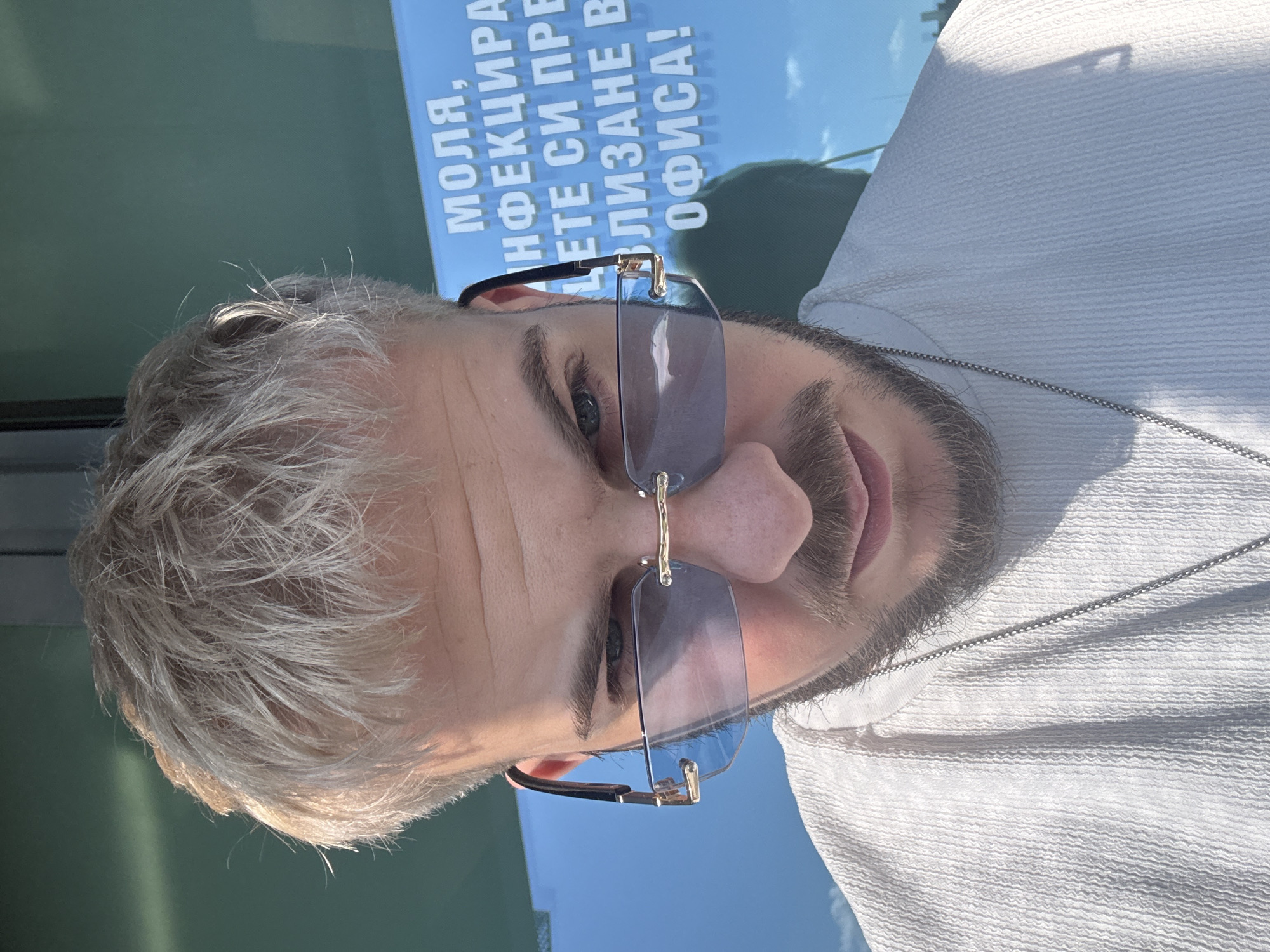 A man with sunglasses, light hair, and a beard is positioned in front of a poster with text in Bulgarian. The text includes the words "МОЛЯ" and "ОФИСА!". The man is wearing a white shirt and a silver chain. The background shows a sky and green surface, suggesting an outdoor location. Given the Bulgarian text, the location is likely Sofia, Bulgaria.