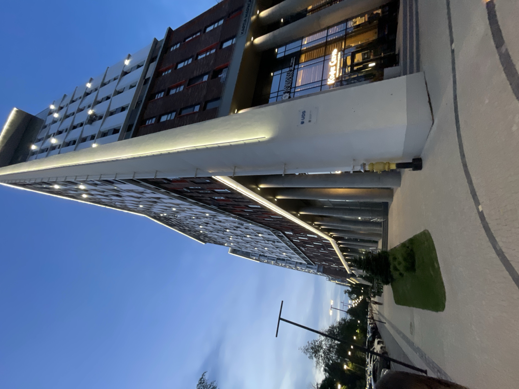 The image shows a modern building in Izhevsk, Russia, captured from a low angle. The building has a combination of white and reddish-brown brick facade with numerous windows. A large white overhang with integrated lighting is visible. There's signage in Russian on the building, possibly indicating "Карта Магика" (Karta Magica) and "Кофе city" (Coffee City). Below the building, there's a paved sidewalk with a strip of green grass and some trees and bushes. A few cars are parked along the curb. The sky is a gradient of blue, indicating either dusk or dawn. A streetlight is also visible in the foreground.