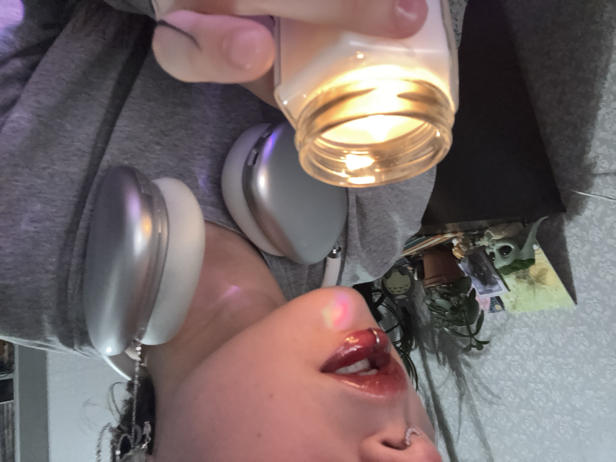 A person is holding a lit candle in a glass jar. They are wearing headphones with metallic silver earcups and a gray hooded sweatshirt. The person has pierced nostrils and is wearing dark, glossy lipstick. In the background, there is a small potted plant, decorative items, and a wallpapered wall. There is no indication that this image was taken in Saint Petersburg, Russia.