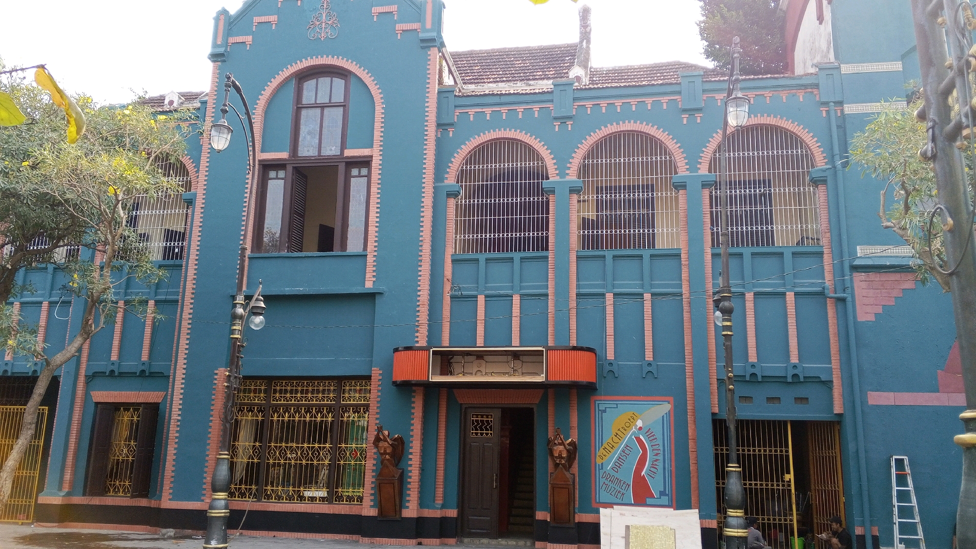 The image shows the facade of an art deco building in Semarang, Indonesia, identified as the Gedung Merdeka (Freedom Building). The building has a distinctive teal-blue exterior with reddish-orange brick accents around arched windows and vertical stripes. The facade features multiple arched windows with white grilles on the upper floors, and a prominent arched entryway on the ground floor. Two wooden statues flank the entryway. To the right of the entrance, a colorful mural with text in Dutch and Indonesian is visible, advertising "Dansen Dranken Muziek" (Dancing Drinks Music) and "Vier den Nacht" (Celebrate the Night). A street lamp stands in front of the building, and trees with yellow flowers are visible on the left. A ladder leans against the right side of the building. Two individuals are partially visible entering or exiting the building through a gated doorway on the right. The sky is overcast.