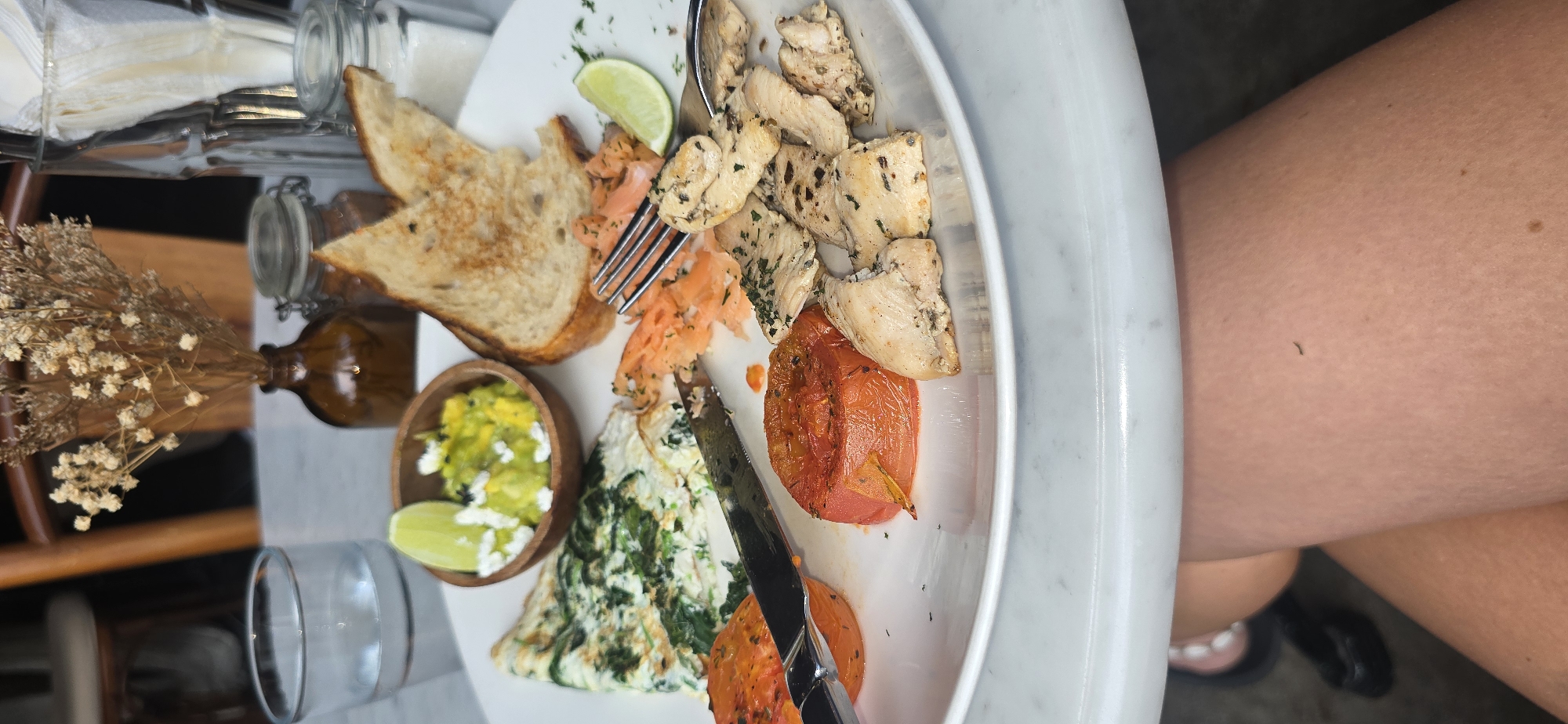A close-up, eye-level shot shows a white plate of food on a marble-topped table. The food consists of an omelet with spinach, a small bowl of guacamole with feta cheese and a lime wedge, two slices of toasted bread, smoked salmon, grilled chicken pieces, and two halves of a roasted tomato. A fork and knife are resting on the plate. In the background, a glass of water, a small brown vase with dried flowers, and a glass jar with a lid are visible. The lower portion of the image shows a person's legs. This scene is consistent with a meal served at a cafe or restaurant in Kuta, Indonesia, given the casual dining setup and the presence of a typical breakfast/brunch spread.
