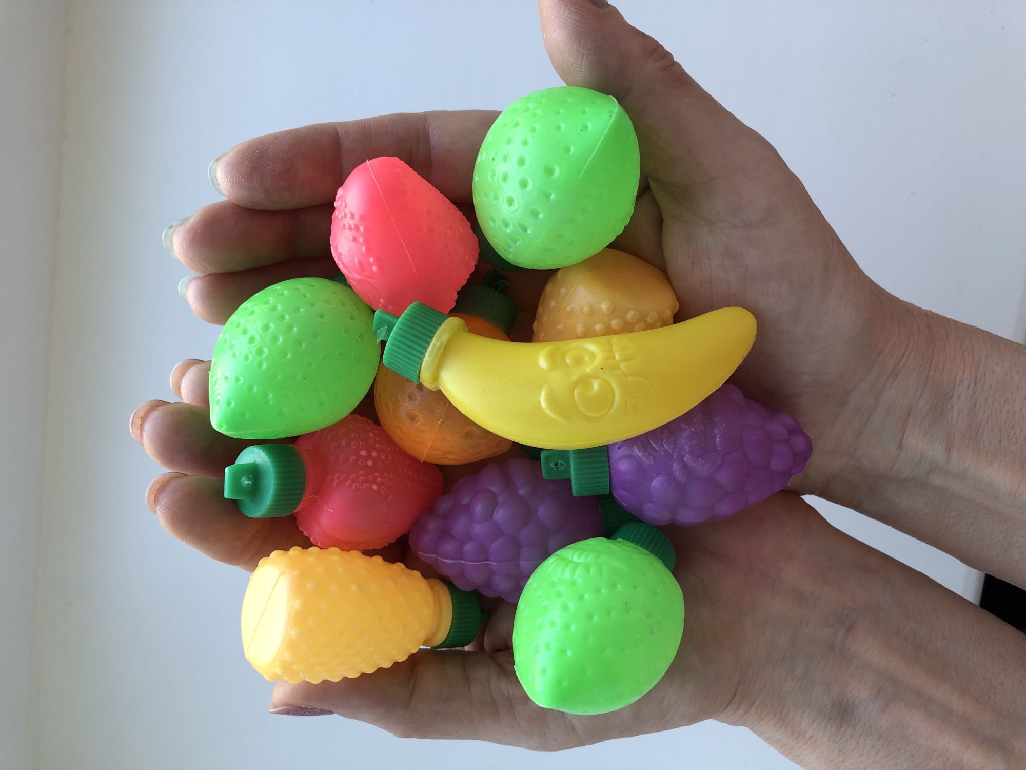 A pair of hands holds a collection of colorful plastic toy fruits. The fruit includes green spheres, a pink strawberry shape, a yellow banana, yellow and orange oblong shapes, and purple grapes. The scene is indoors, with a white background. The location is not discernible from the image.