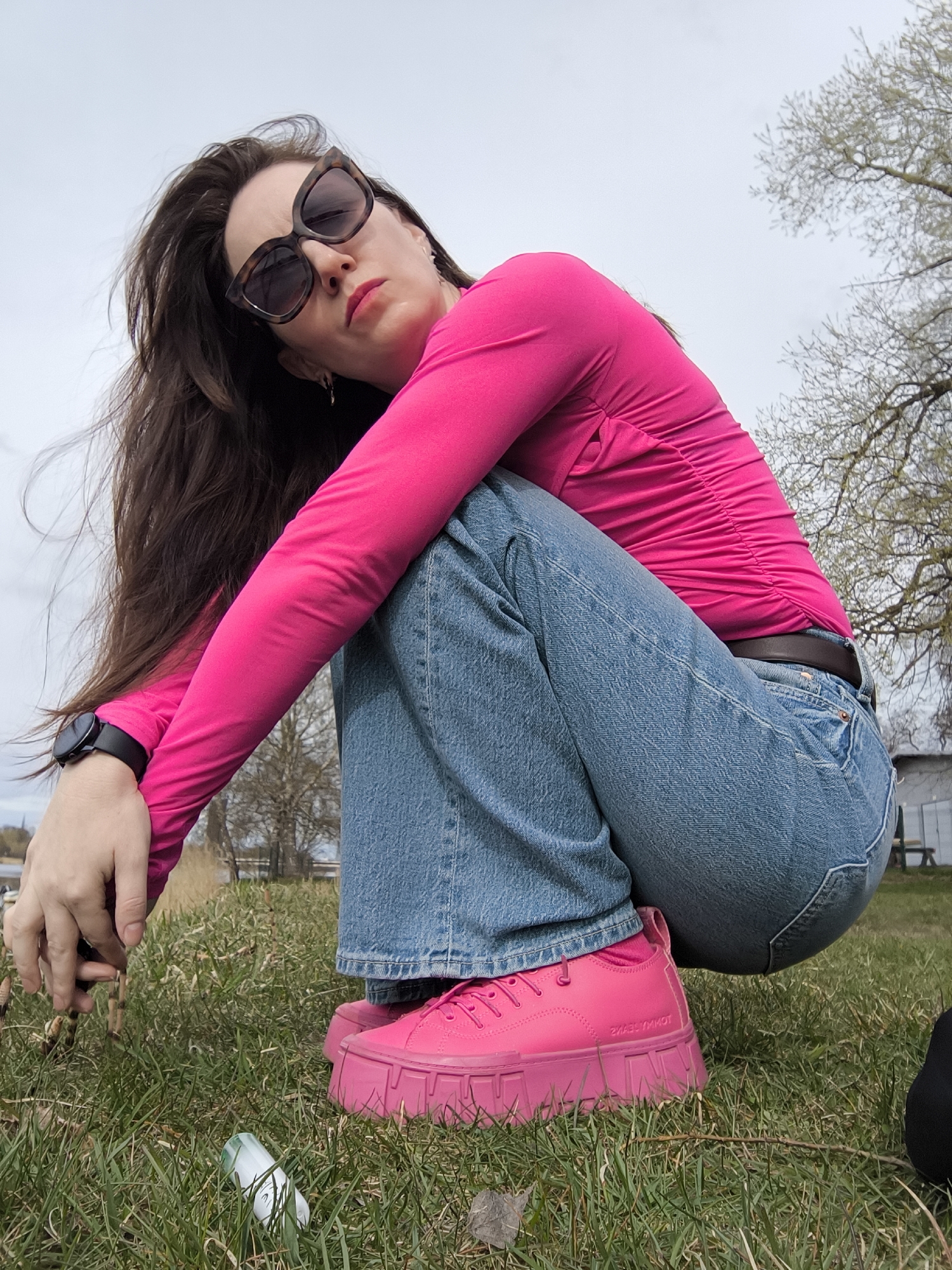 A woman is squatting on a grassy area outdoors. She wears a bright pink long-sleeved shirt, blue jeans, and bright pink platform sneakers. She also wears dark sunglasses and a watch on her left wrist. Her long brown hair is blowing in the wind.
In the foreground, there is grass and a white plastic container lying on the ground. In the background, there are trees with bare branches and a body of water. A building is visible in the distance.
While there is no explicit information confirming that this photo was taken in Riga, Latvia, the presence of features of the environment is not enough to conclude that the photo was taken in the city of Riga, Latvia.