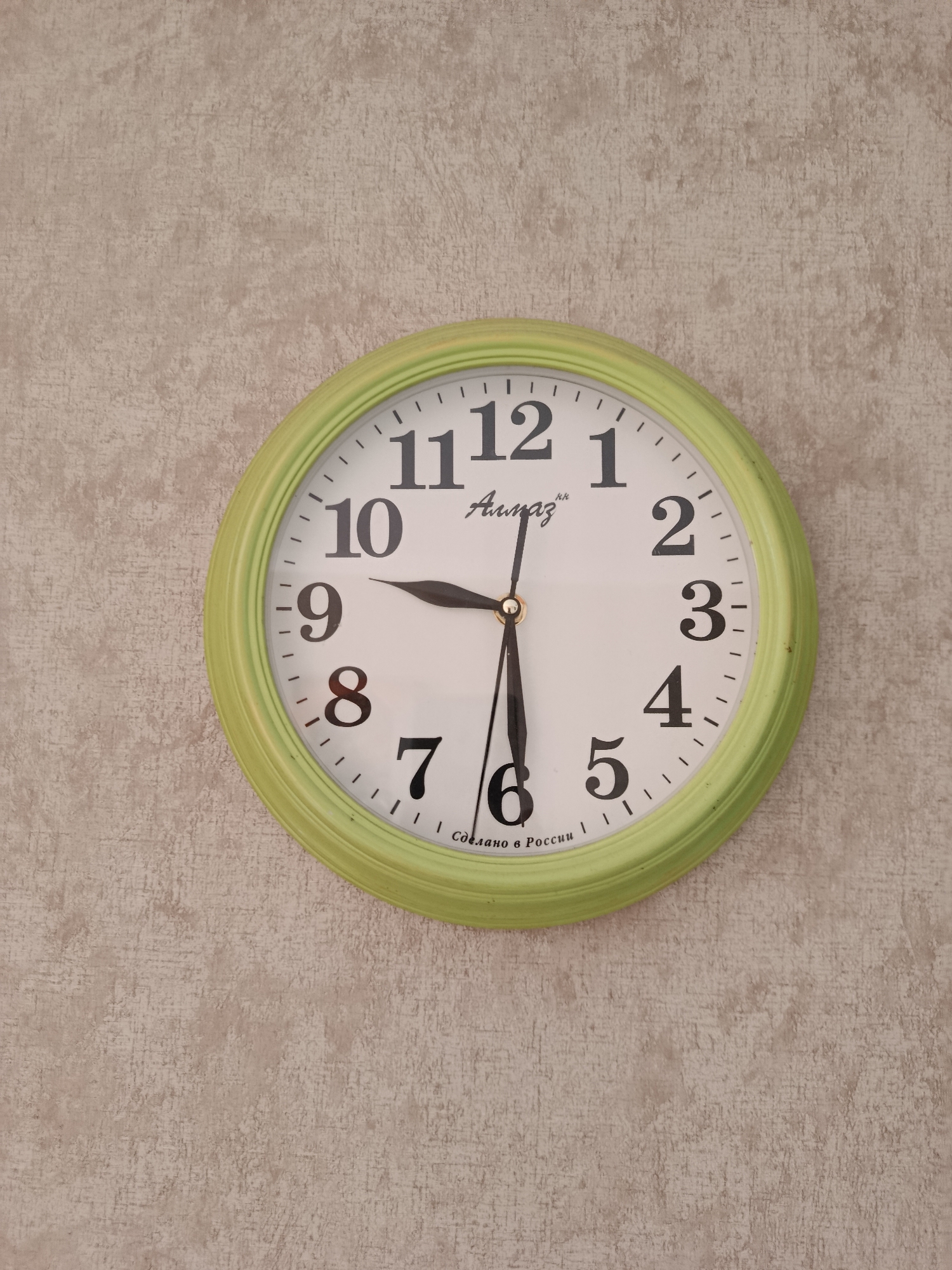 A light green analog wall clock is affixed to a beige, textured wall. The clock face is white with black Arabic numerals marking the hours from 1 to 12. The hour hand is pointing to the 9, and the minute hand is pointing to the 6. The second hand is a thin black line and is also pointing to the 6. The brand name "Алмаз" (Almaz) is visible above the center of the clock face. At the bottom of the clock face, it reads "СДЕЛАНО В РОССИИ" (Made in Russia). The clock is the sole subject of the image. The wall appears to be an interior wall, possibly in a residential or office setting, in the city of Biysk, Russia.