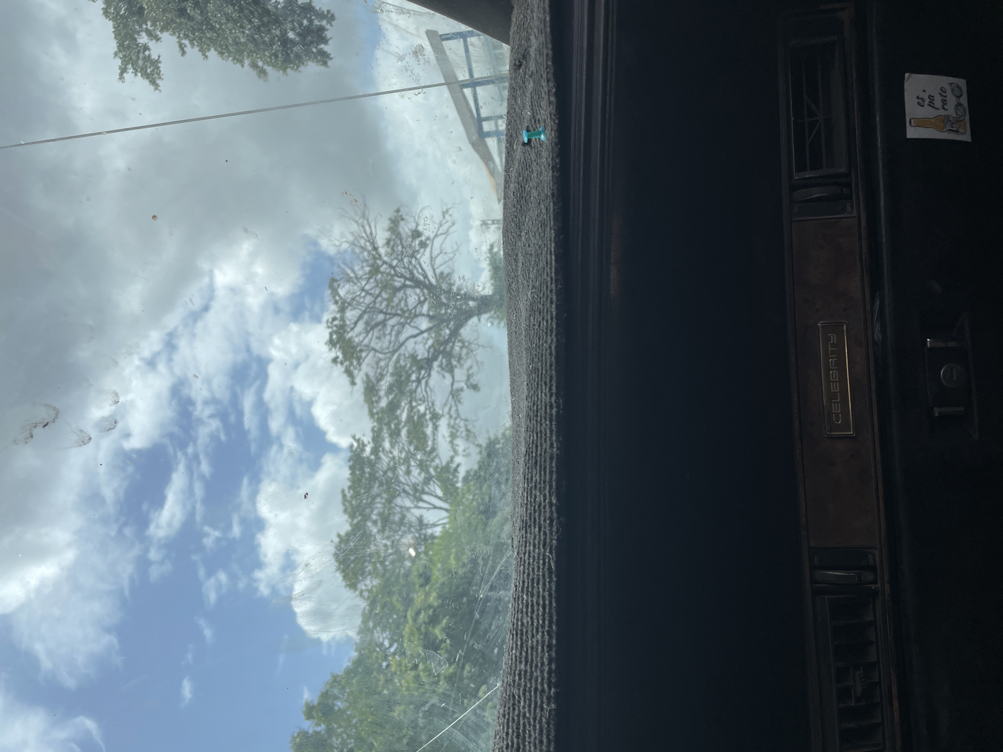The image is a view from inside a vehicle looking out through the windshield. The windshield is dirty and shows smudges. Outside, a cloudy sky with patches of blue is visible, along with green trees and a structure that appears to be part of a playground or park. The dashboard of the car is visible in the foreground, with an air vent and the word "CELEBRITY" embossed on a metallic panel. A sticker with an illustration of a beer bottle and glasses, and text that reads "es, pa rato" is present on the lower right side of the dashboard. The scene is likely in Juan Griego, Venezuela, based on the context provided.