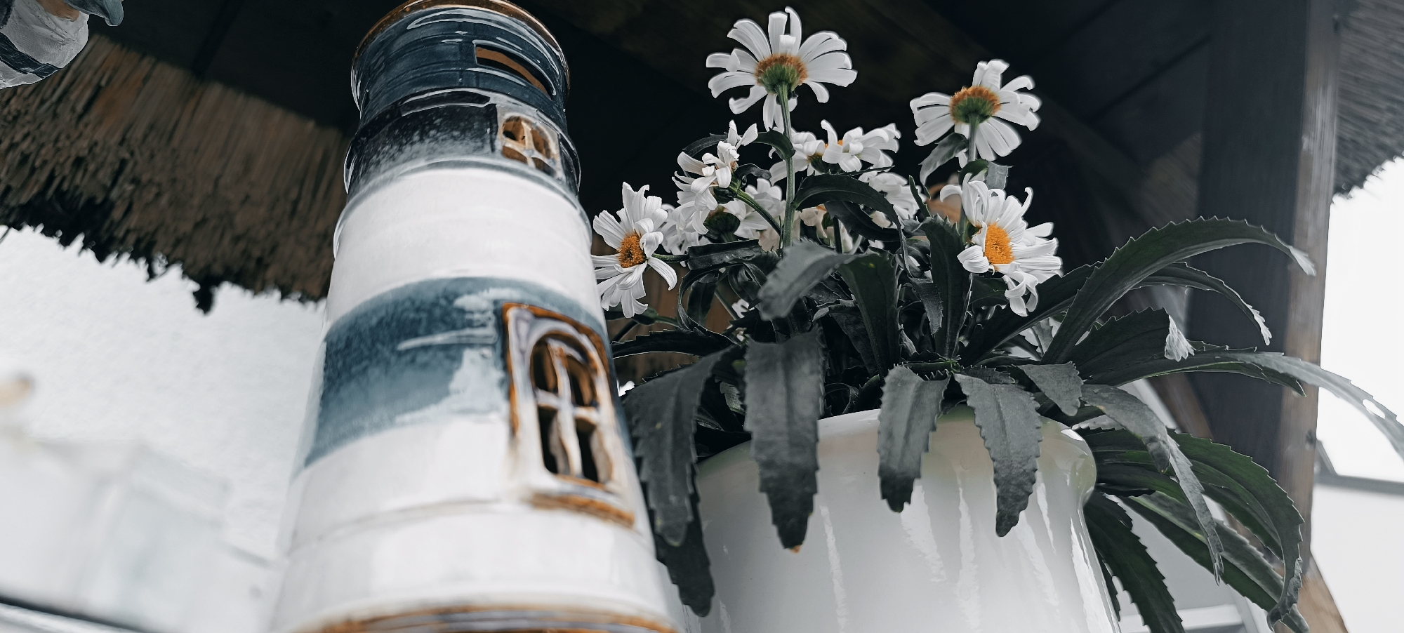 The image shows a close-up of a decorative ceramic lighthouse next to a white pot containing white daisies. The lighthouse is white with blue accents and has a window with a brown frame. The daisies have yellow centers and are in full bloom, with dark green leaves. The background is blurred, showing a thatched roof and some wooden beams, suggesting an outdoor setting, possibly a garden or patio. The overall mood is peaceful and decorative. This image was taken in Waiblingen, Germany.