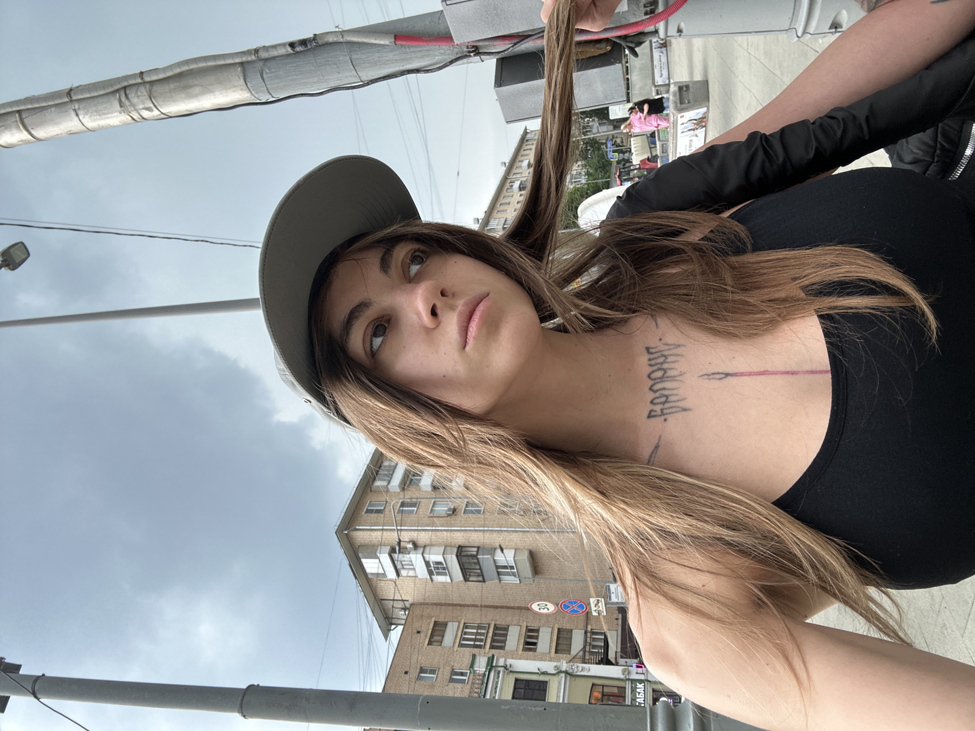 A young woman with long, light brown hair, wearing a grey baseball cap and a black tank top, poses for a selfie in an outdoor urban setting. She has a tattoo on her chest and is holding a lock of her hair. In the background, there are multi-story brick buildings, street signs, power lines, and a lamppost. The sky is overcast. The scene appears to be a street in Moscow, Russia, as indicated by a traffic sign displaying "30" which is a common speed limit in Russian cities.