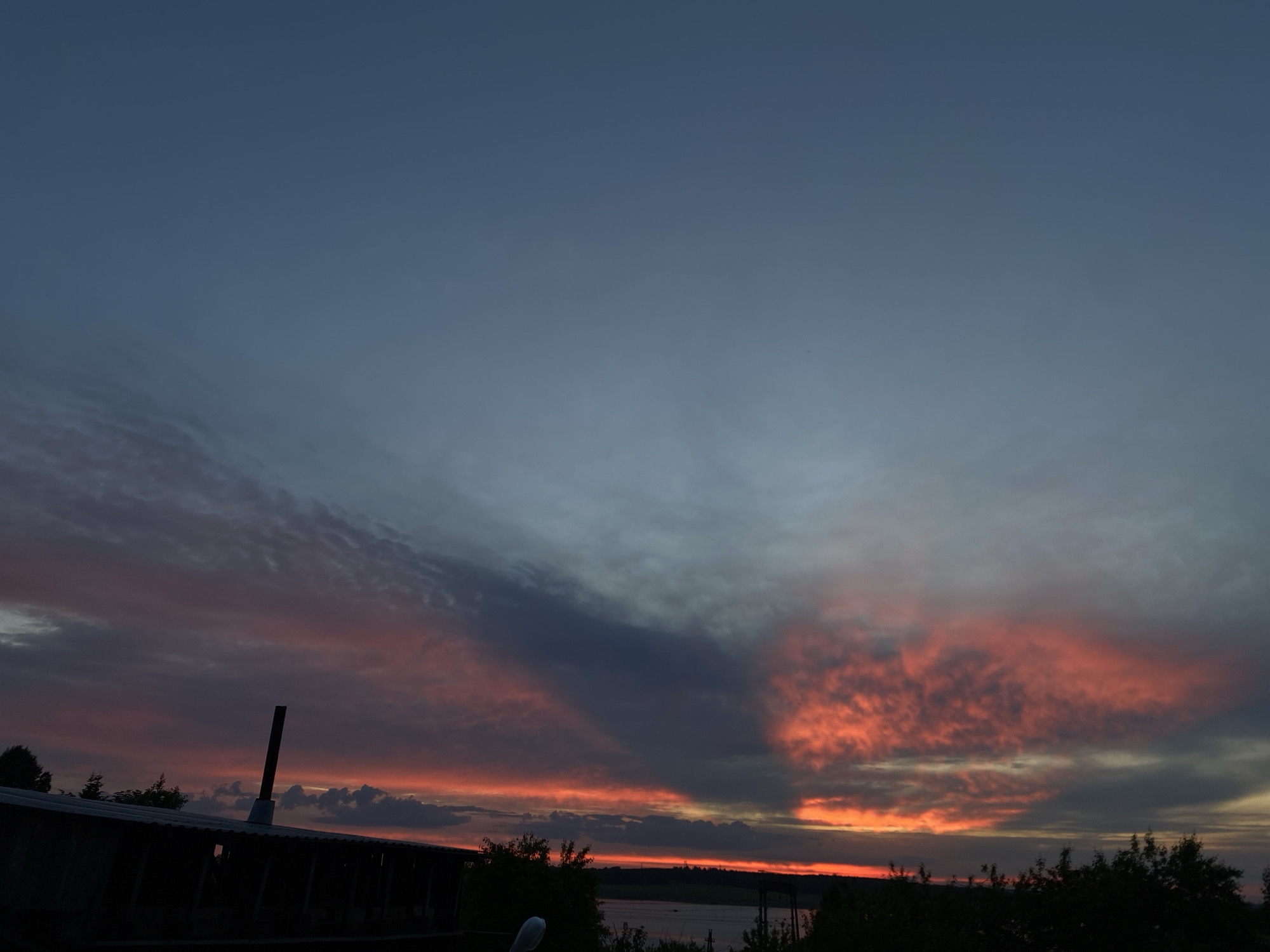 The image presents a dramatic sunset or sunrise over a rural or lakeside landscape in Smolino, Russia. The sky dominates the frame, showcasing a striking gradient from deep blue-grey at the top to vivid reds and oranges closer to the horizon. Numerous clouds are visible, some appearing as delicate, streaky formations, while others form denser, darker patches. A notable dark, fan-shaped cloud formation extends from the lower right towards the center-left, creating a strong contrast with the brightly illuminated areas of the sky.
In the foreground, silhouetted against the colorful horizon, is the roofline of a building, possibly a shed or barn, with a tall, dark pipe or chimney rising from it on the left side. Below the vibrant sky, a thin strip of water, likely a lake or river, is visible in the distance, bordered by the dark silhouettes of trees and vegetation along the horizon and mid-ground. No people or visible text are present. The scene conveys a sense of peaceful twilight, highlighting the natural beauty of the sky and the serene atmosphere of the location.