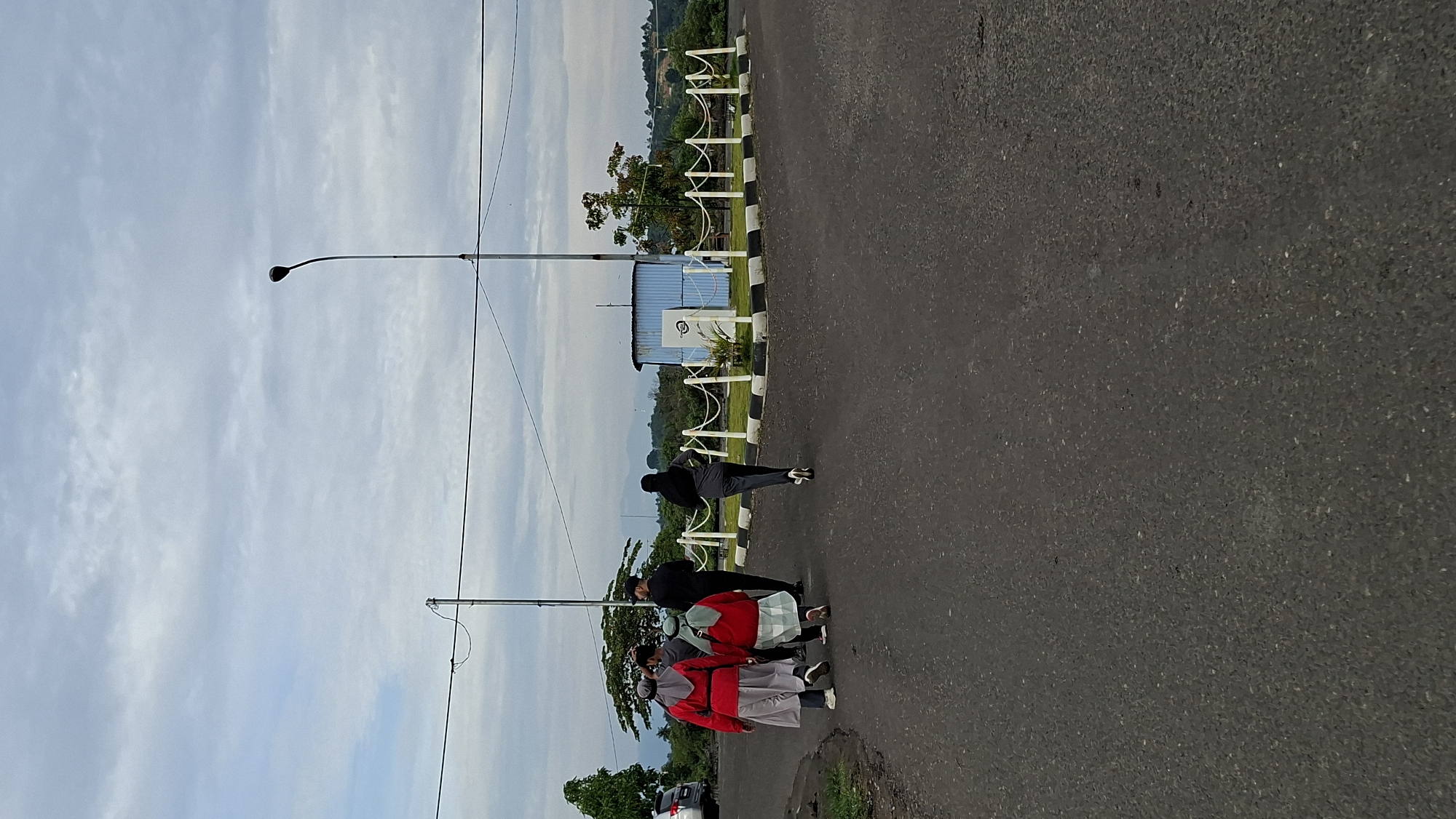 In Lhokseumawe, Indonesia, on an overcast day, four individuals walk away from the camera on a paved road. The group consists of two women in red tops and light-colored skirts or pants, one woman in a gray tracksuit and black hijab, and one person in a black top and dark pants. They are walking towards a small, light blue, shed-like structure with a white railing in front of it. The shed has a sign on its exterior. Behind the shed, trees and distant hills are visible. A tall streetlamp stands to the right of the shed, and power lines cross the sky above. A white vehicle is parked on the left side of the road in the background. The overall environment appears to be an open area with sparse vegetation and infrastructure.