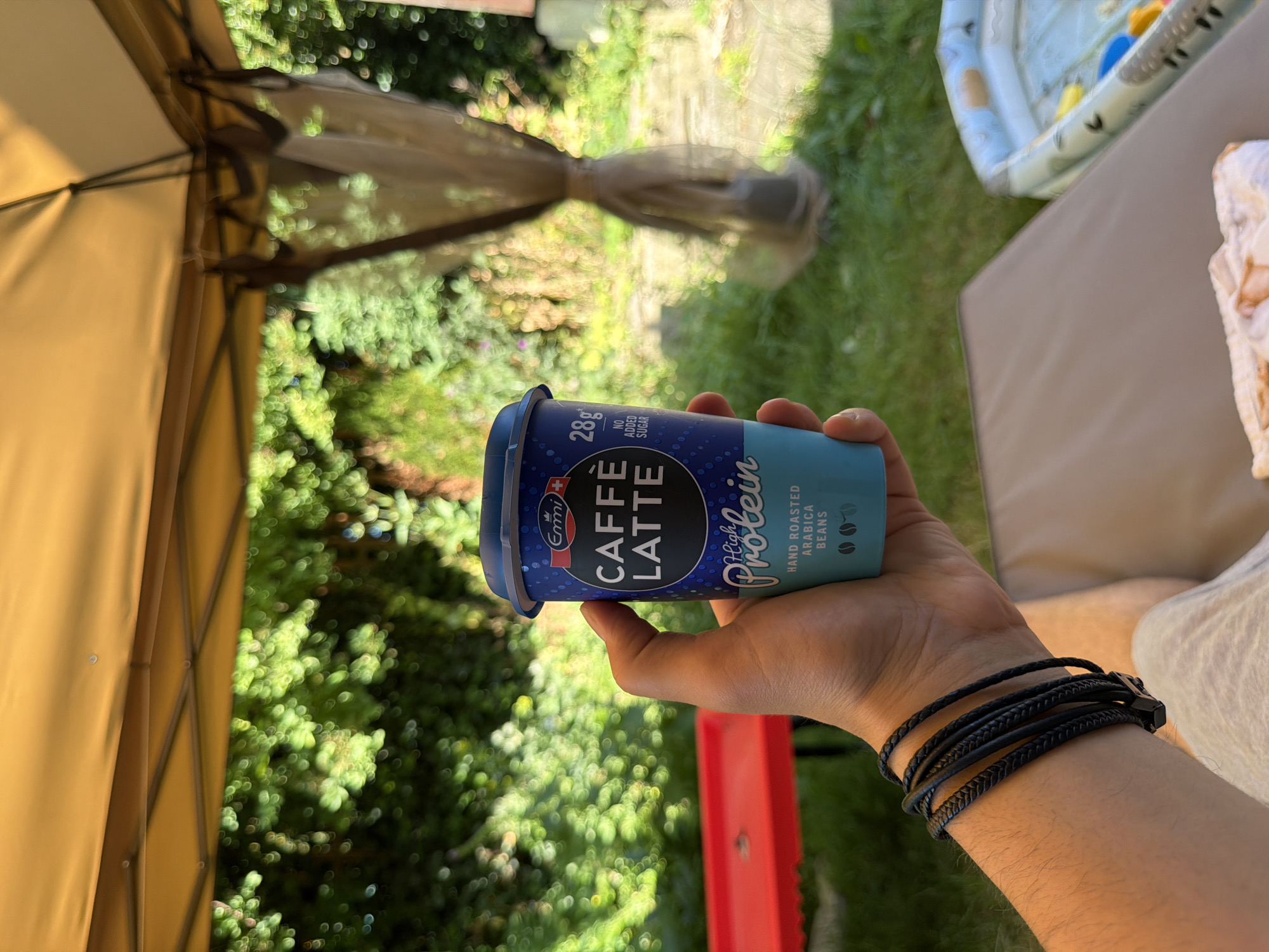A person's hand holds a blue and teal colored Emmi Caffè Latte High Protein drink in a cardboard cup. The cup has white text on a dark blue circular label that says "Emmi CAFFÈ LATTE" and "High Protein". Below this, the text "HAND ROASTED ARABICA BEANS" is visible, along with three small coffee bean icons. A small "28g" is also visible on the cup, with "NO ADDED SUGAR" below it. The hand also wears a black braided leather bracelet. The background is a garden scene with a yellow canopy overhead, green foliage, a red tray on the left, and a blue inflatable pool on the right. The setting is a garden which may be located in Leeds, United Kingdom.