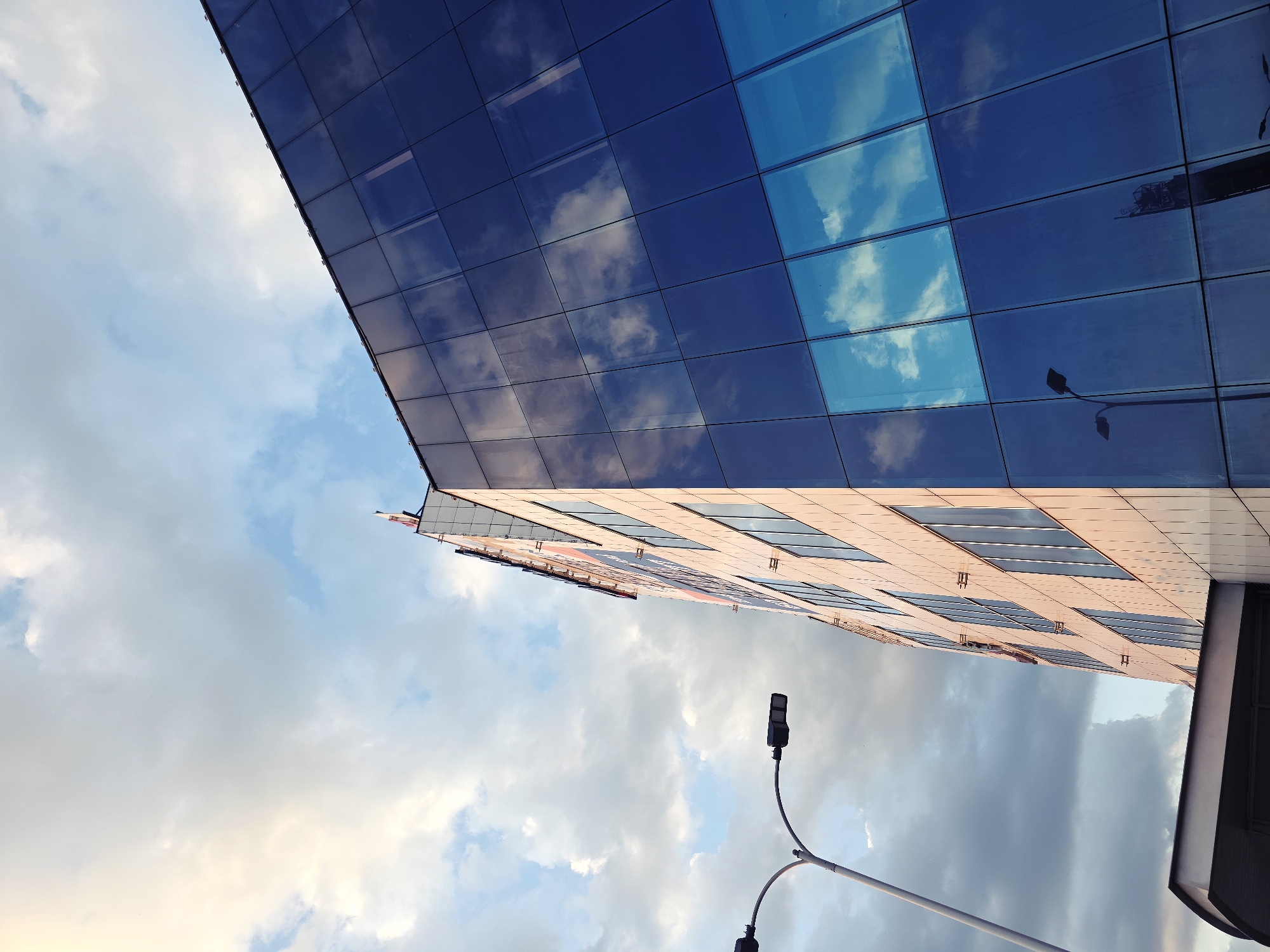 The image depicts the exterior of a modern building in Warsaw, Poland, with a striking glass facade reflecting the sky and clouds. The building's design features a grid of blue glass panels that mirror the sky's hues, creating a visual interplay between the structure and its environment. The lower portion of the building appears to be constructed with a lighter, possibly cream-colored material, also arranged in a grid pattern. A street lamp with two lamps extends into the foreground. The sky is filled with clouds.